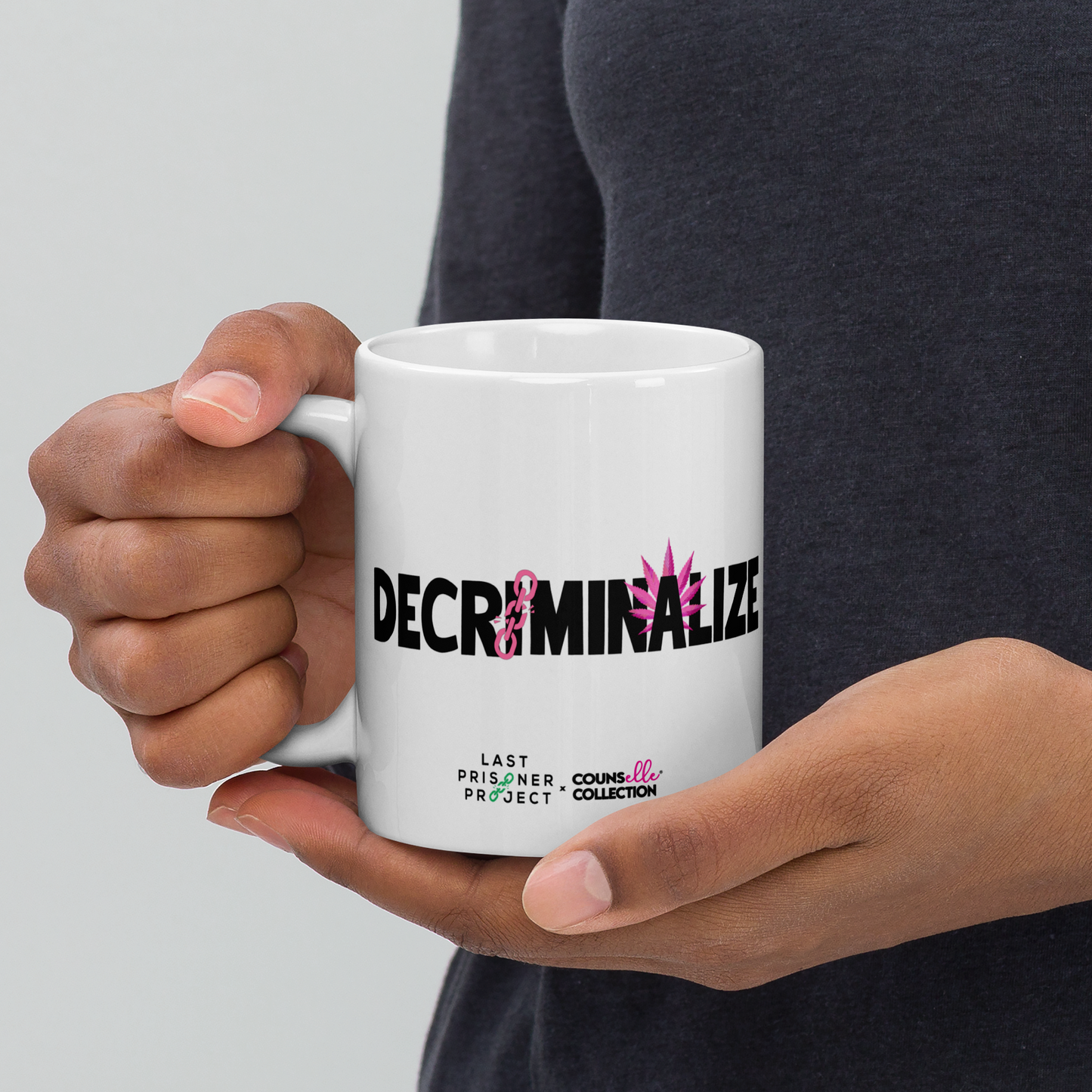 A person in a dark grey shirt holding a white mug with the text "decriminalize" on the front in their hands.
Last Prisoner Project and Counselle Collection® logos at the bottom of the mugs.
