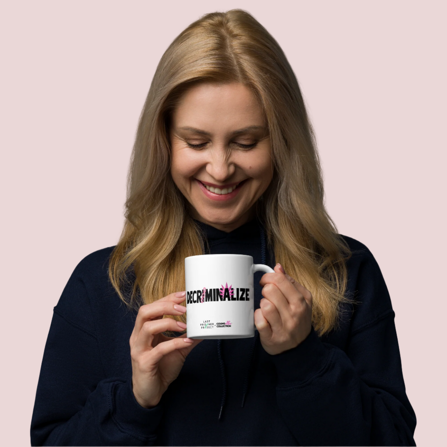A white woman with mid-length blonde hair wearing a navy blue long sleeved hoodie holding a white mug with the text "decriminalize" on the front. The woman is looking down and smiling.
Last Prisoner Project and Counselle Collection® logos at the bottom of the mugs.