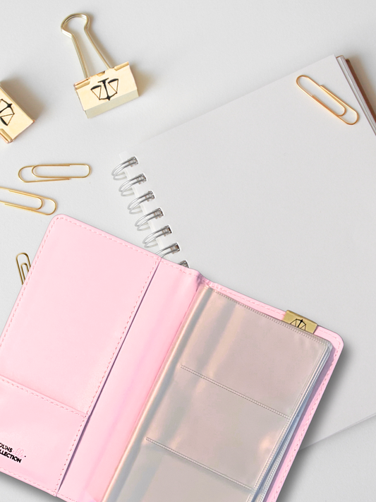 a vegan leather pink business card holder on a white background surrounded by gold paper and binder clips and a white spiral notebook