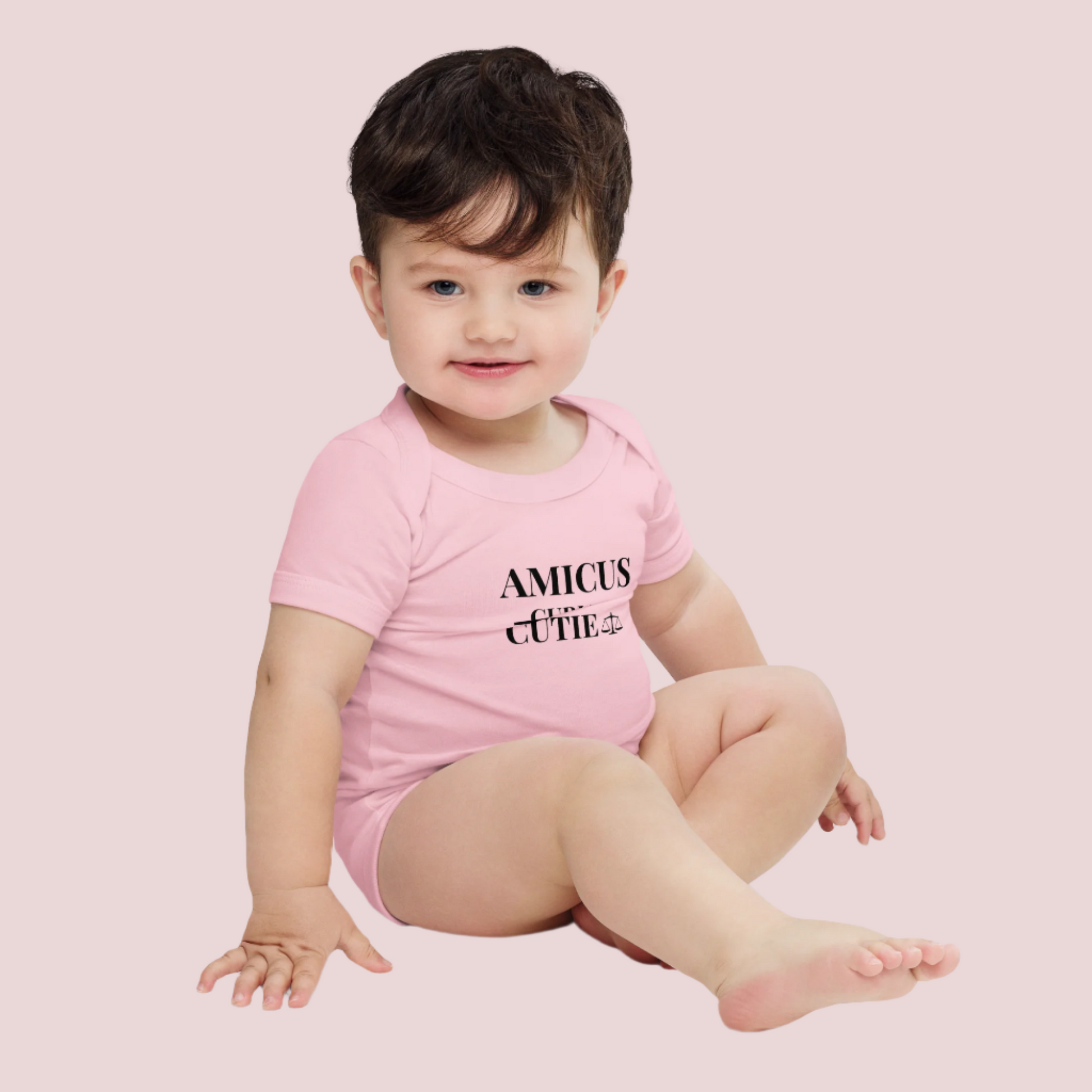 An infant wearing a pink onesie featuring in black text, the words "AMICUS", "CUTIE", and a stricken out word "CURIAE", with an illustration of justice scales.