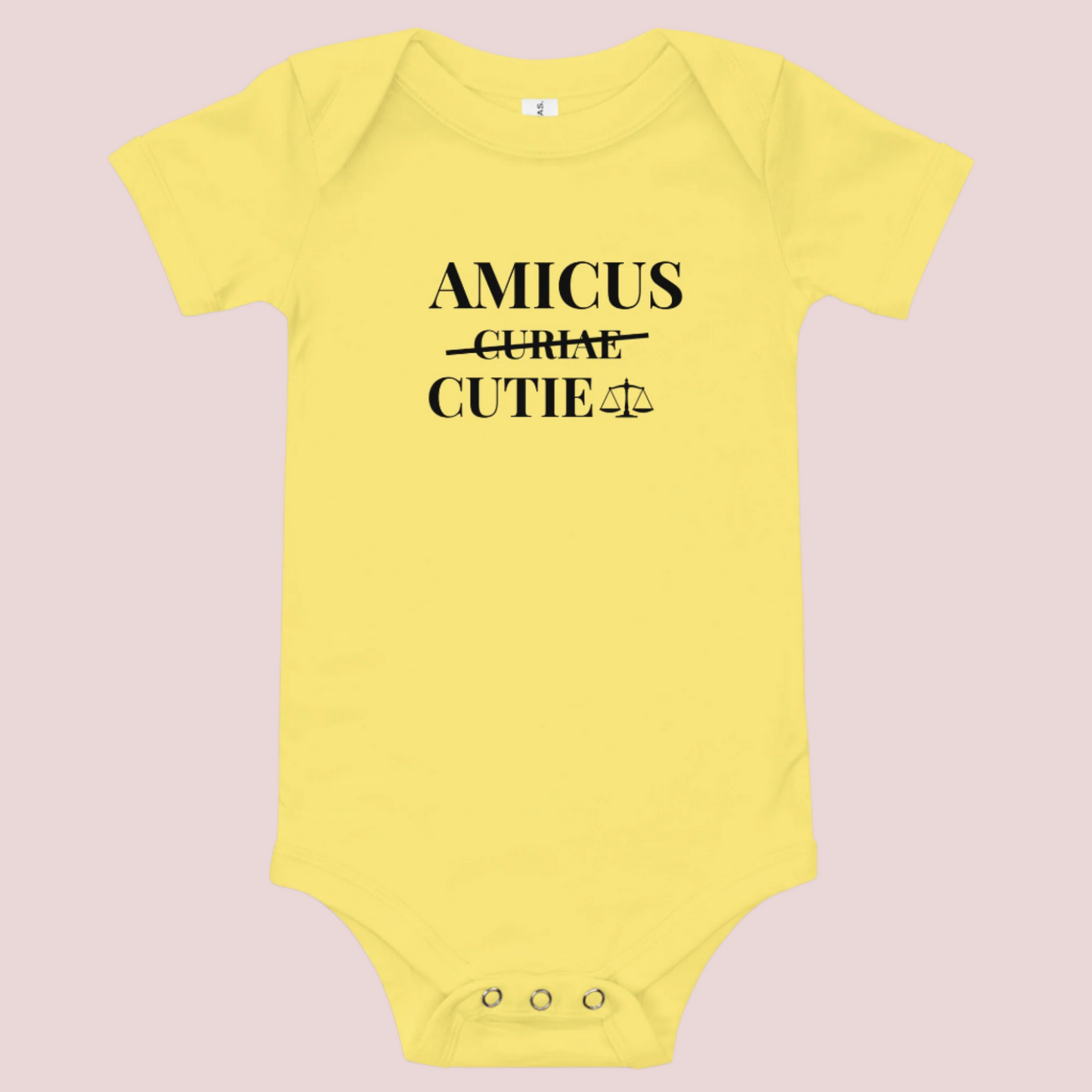 A yellow infant onesie featuring in black text, the words "AMICUS", "CUTIE", and a stricken out word "CURIAE", with an illustration of justice scales.