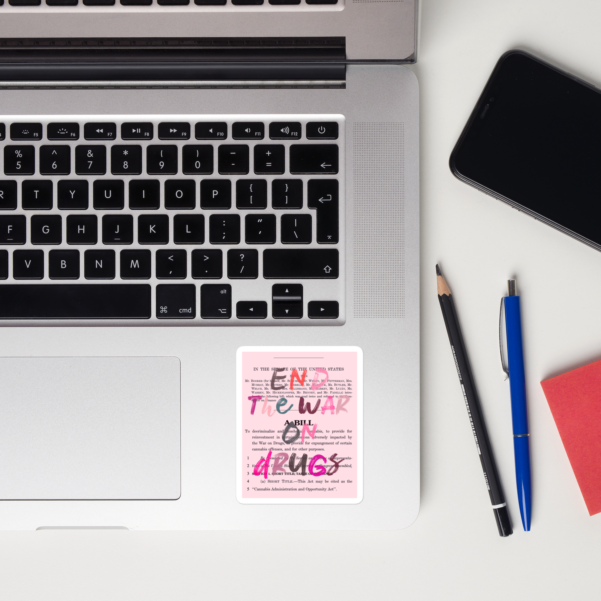 A sticker showing the text "End The War on Drugs" written in different colored font,  placed on a laptop. A pen, pencil, notepad, and mobile phone can also be seen on the flatlay.