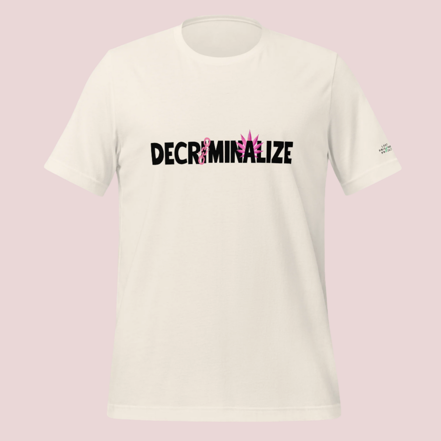 A cream crewneck tee showing the text "DECRIMINALIZE", with a broken pink chain over the first letter "I" and pink cannabis leaves behind the letter "A" . The Last Prisoner Project x The Counselle Collection logo on the left sleeve.