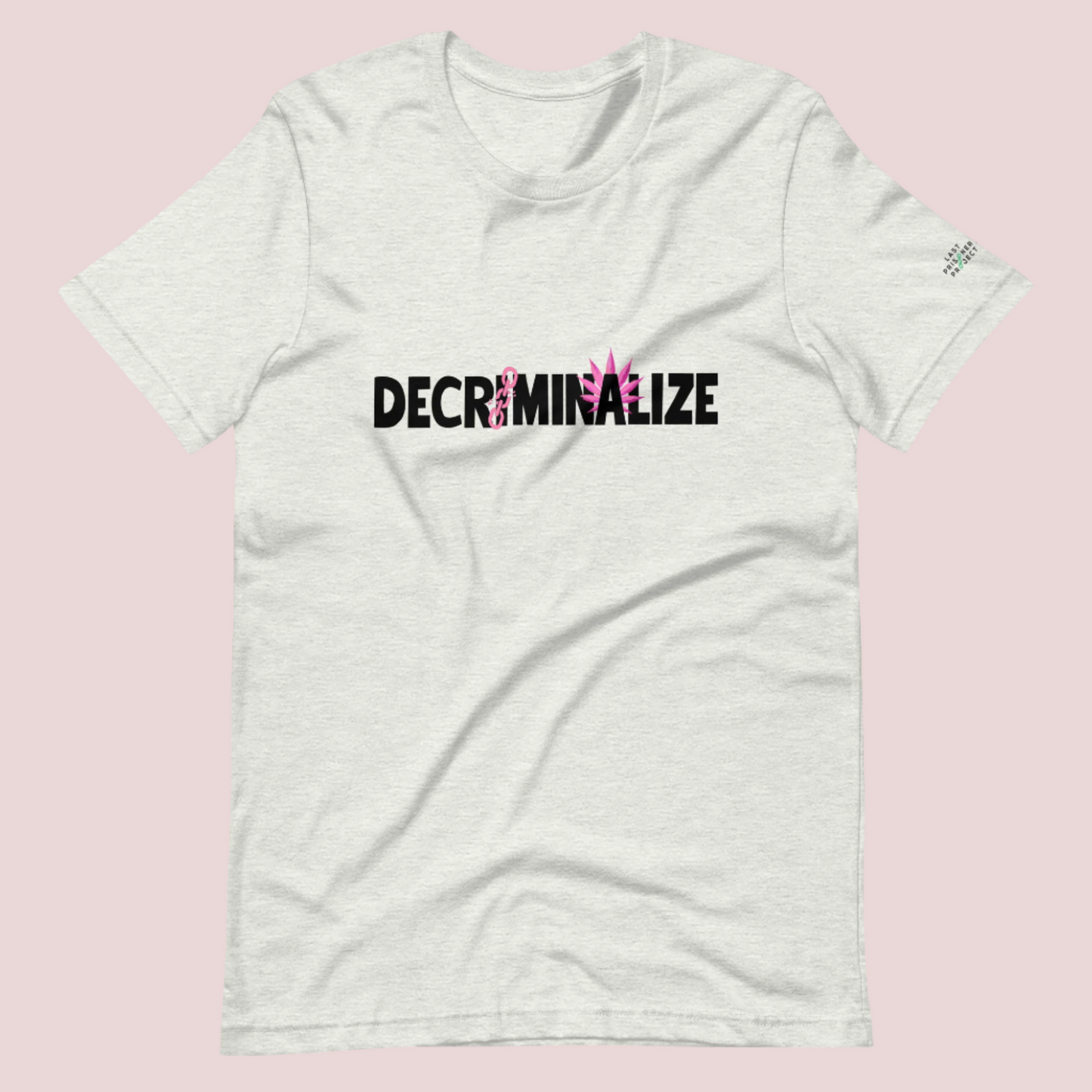 A grey crewneck tee showing the text "DECRIMINALIZE", with a broken pink chain over the first letter "I" and pink cannabis leaves behind the letter "A" . The Last Prisoner Project x The Counselle Collection logo on the left sleeve.