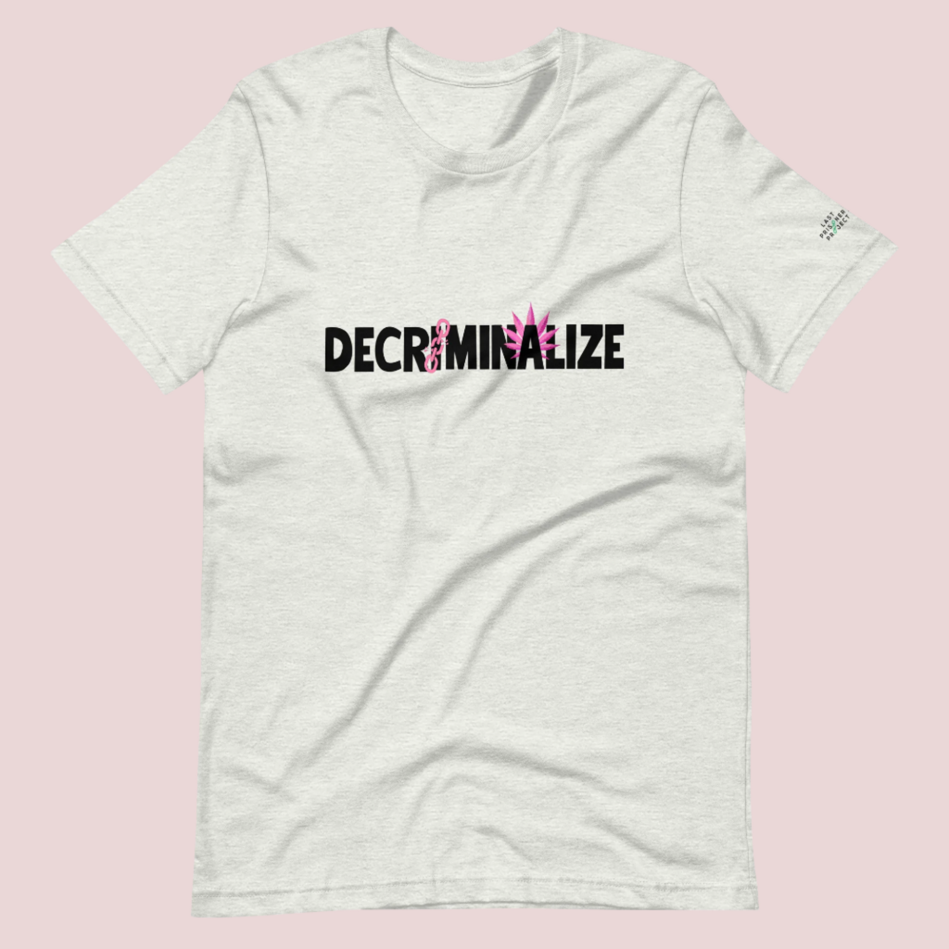 A grey crewneck tee showing the text "DECRIMINALIZE", with a broken pink chain over the first letter "I" and pink cannabis leaves behind the letter "A" . The Last Prisoner Project x The Counselle Collection logo on the left sleeve.