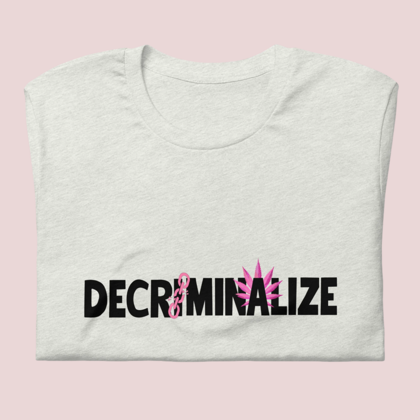 A folded grey crewneck tee showing the text "DECRIMINALIZE", with a broken pink chain over the first letter "I" and pink cannabis leaves behind the letter "A" .