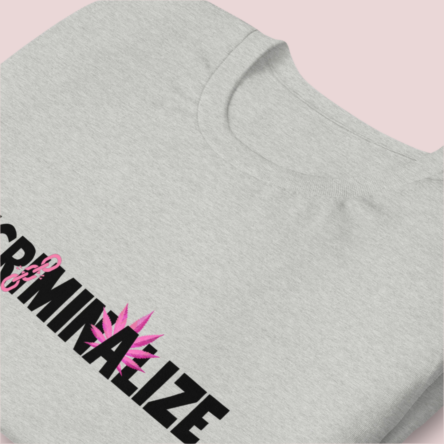 A folded grey crewneck tee showing the text "DECRIMINALIZE", with a broken pink chain over the first letter "I" and pink cannabis leaves behind the letter "A" .