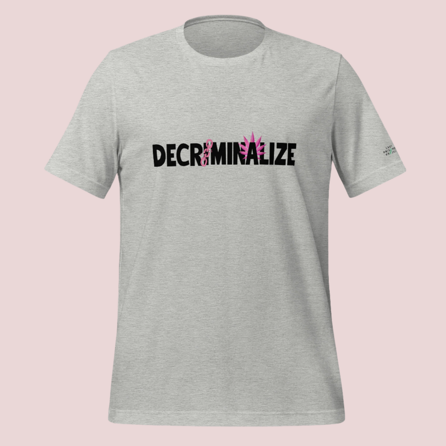 A grey crewneck tee showing the text "DECRIMINALIZE", with a broken pink chain over the first letter "I" and pink cannabis leaves behind the letter "A" . The Last Prisoner Project x The Counselle Collection logo on the left sleeve.