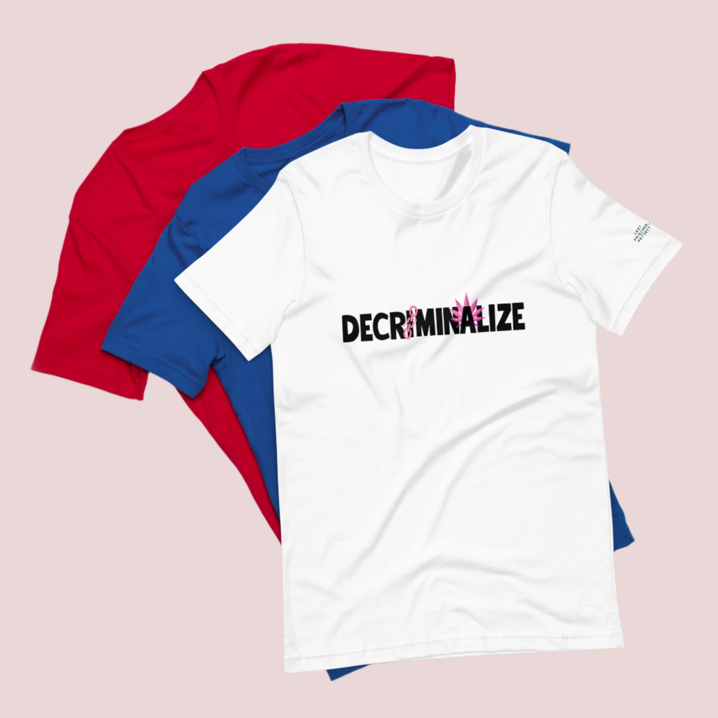 Three crewneck tees, colored red, blue, and white, the white tee showing the text "DECRIMINALIZE", with a broken pink chain over the first letter "I" and pink cannabis leaves behind the letter "A" . The Last Prisoner Project x The Counselle Collection logo on the left sleeve.