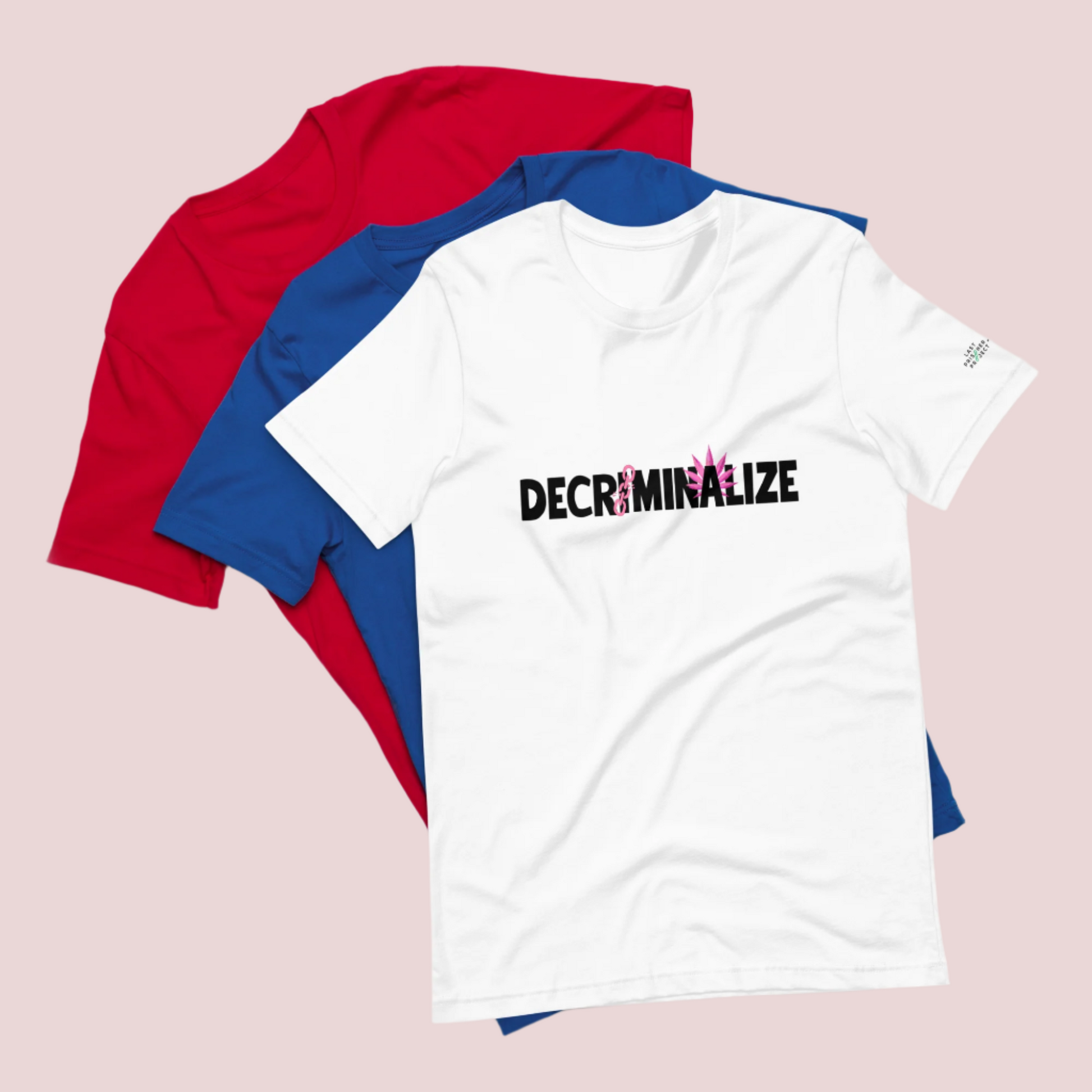 Three crewneck tees, colored red, blue, and white, the white tee showing the text "DECRIMINALIZE", with a broken pink chain over the first letter "I" and pink cannabis leaves behind the letter "A" . The Last Prisoner Project x The Counselle Collection logo on the left sleeve.