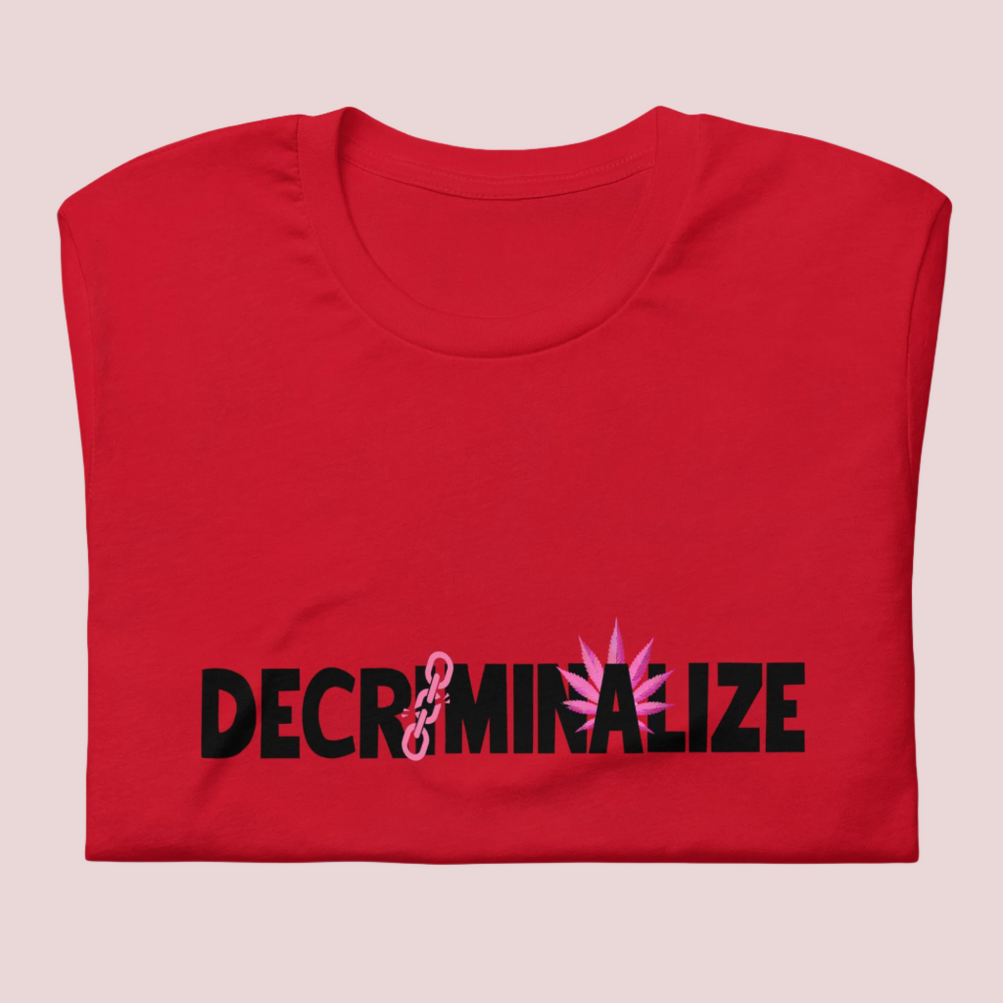 A folded red crewneck tee showing the text "DECRIMINALIZE", with a broken pink chain over the first letter "I" and pink cannabis leaves behind the letter "A" .