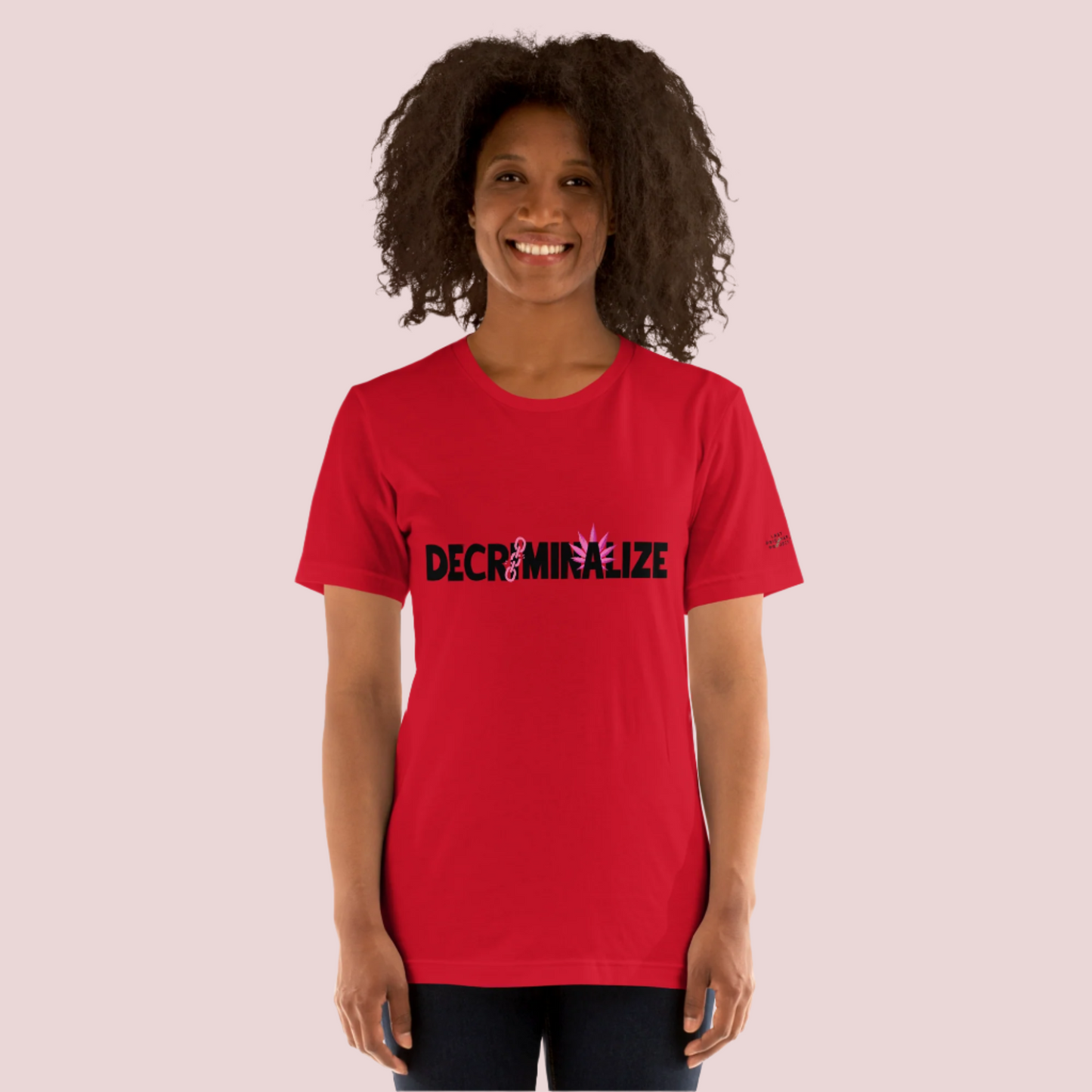 A person wearing a red crewneck tee showing the text "DECRIMINALIZE", with a broken pink chain over the first letter "I" and pink cannabis leaves behind the letter "A" . The Last Prisoner Project x The Counselle Collection logo on the left sleeve.