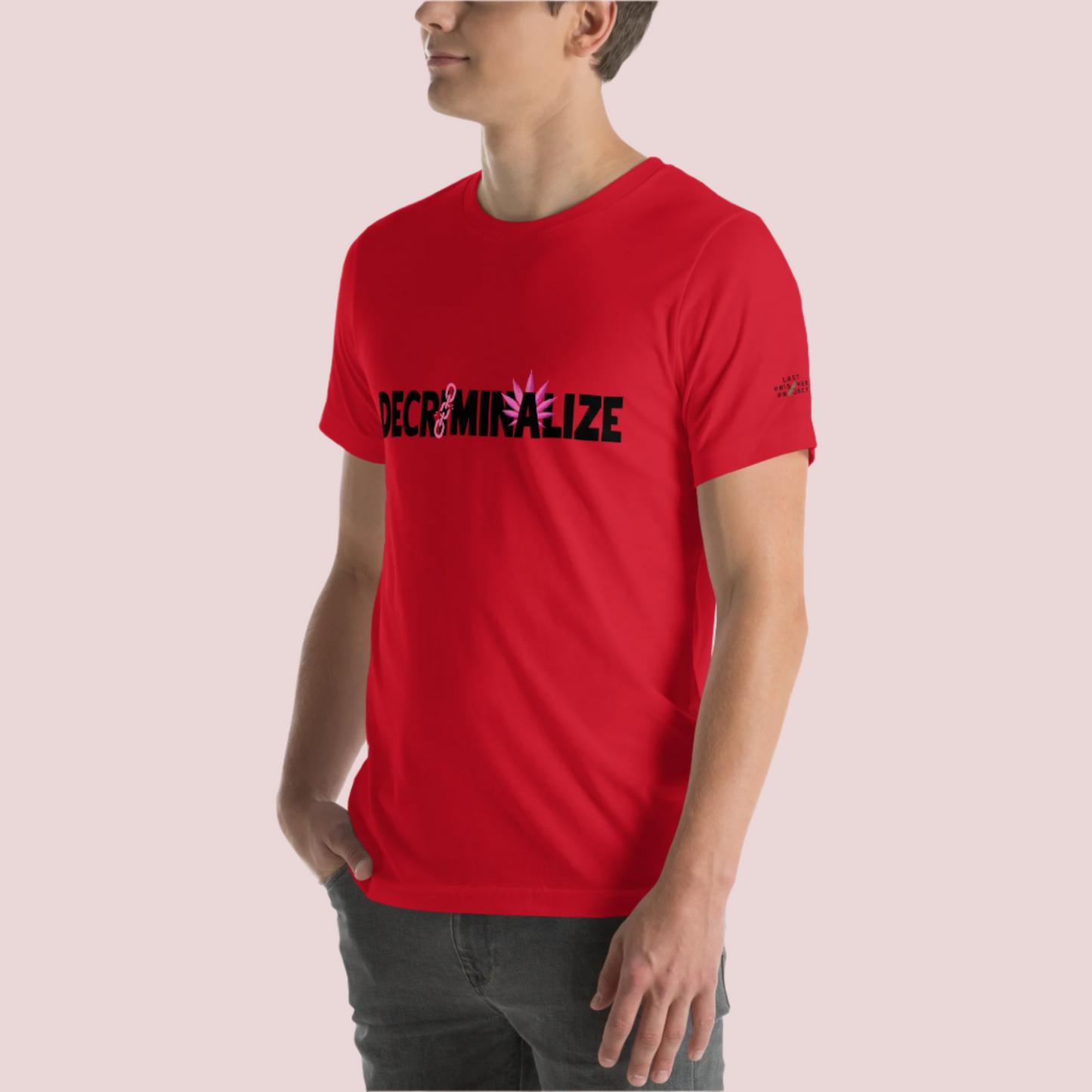 A person wearing a red crewneck tee showing the text "DECRIMINALIZE", with a broken pink chain over the first letter "I" and pink cannabis leaves behind the letter "A" . The Last Prisoner Project x The Counselle Collection logo on the left sleeve.