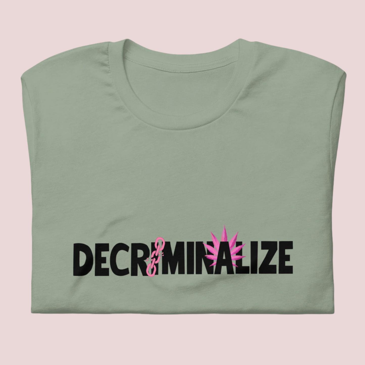 A sage green crewneck tee showing the text "DECRIMINALIZE", with a broken pink chain over the first letter "I" and pink cannabis leaves behind the letter "A" .