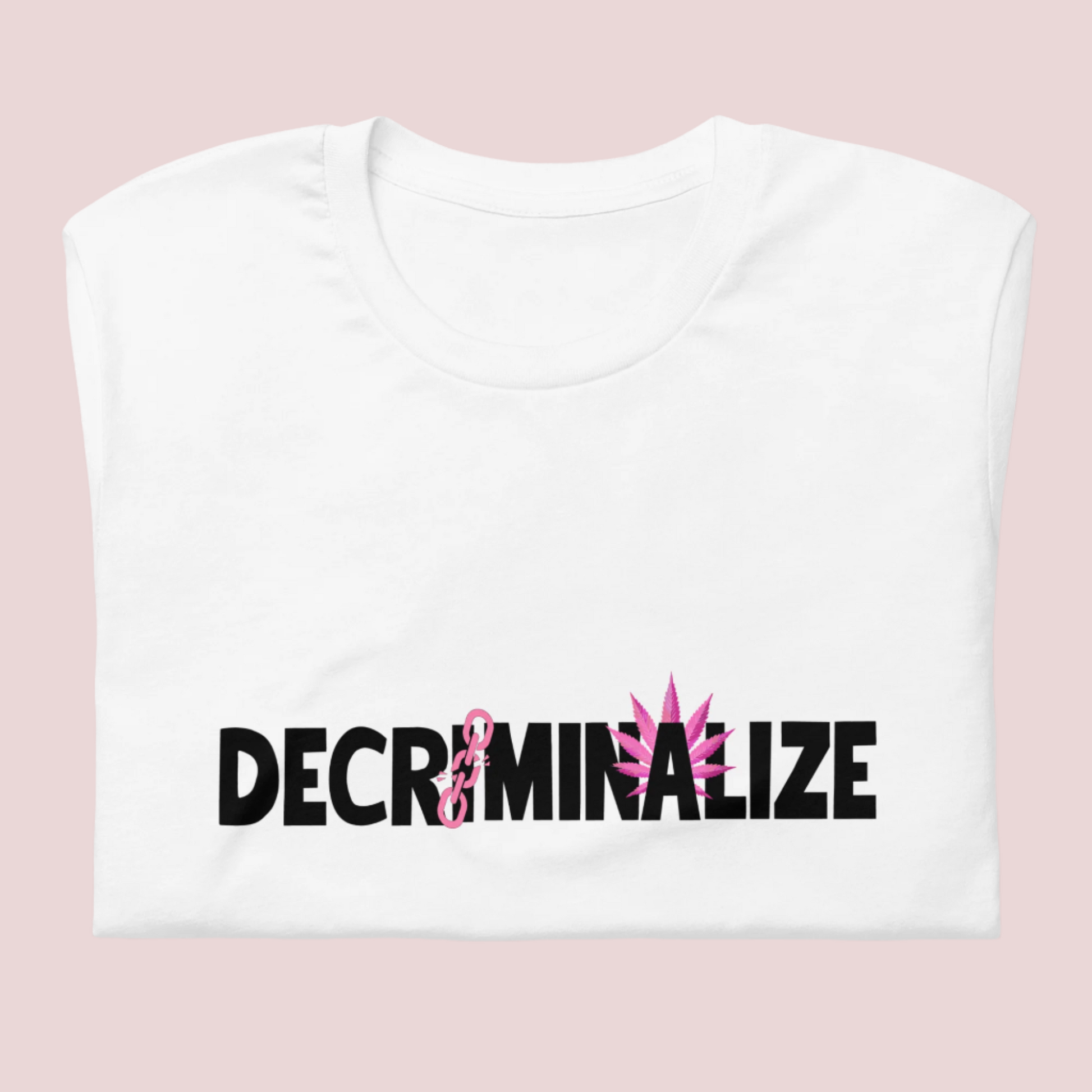 A folded white crewneck tee showing the text "DECRIMINALIZE", with a broken pink chain over the first letter "I" and pink cannabis leaves behind the letter "A" .