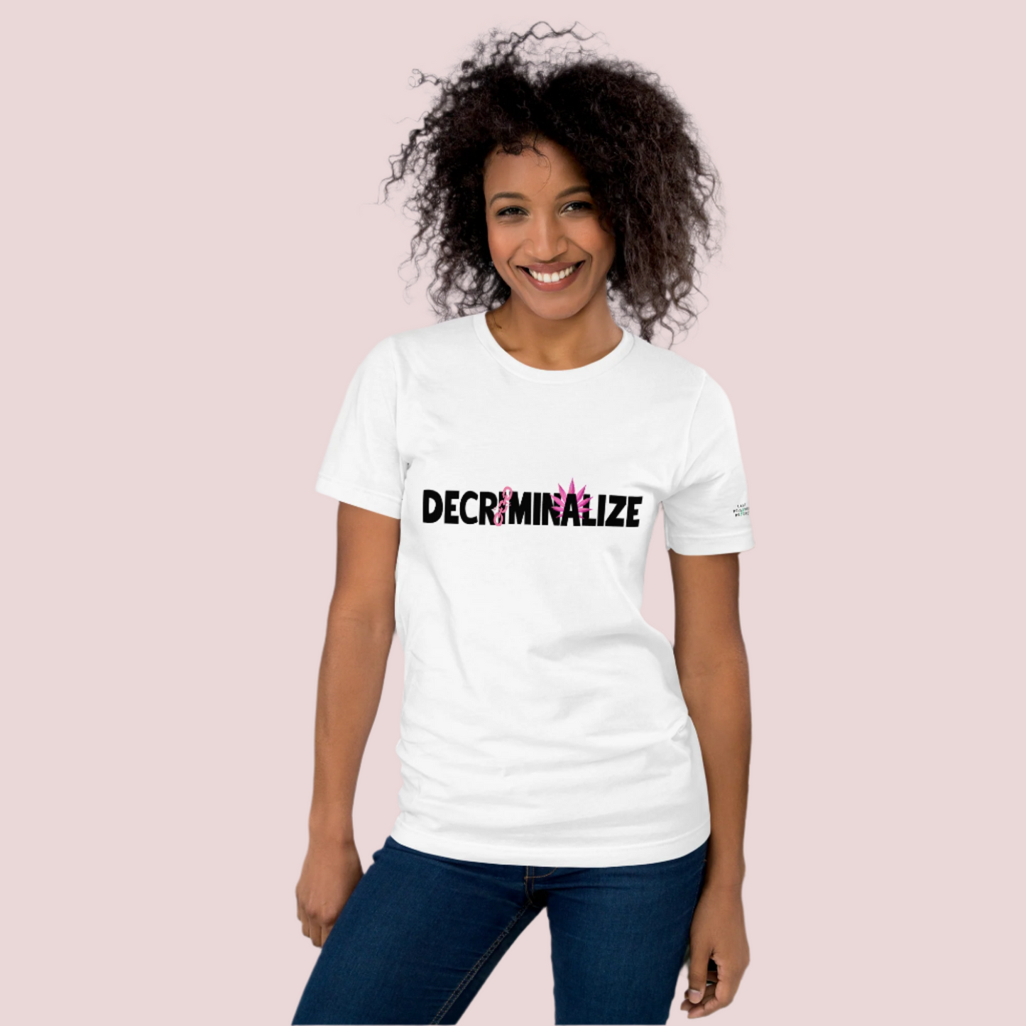 A person wearing a white crewneck tee showing the text "DECRIMINALIZE", with a broken pink chain over the first letter "I" and pink cannabis leaves behind the letter "A" . The Last Prisoner Project x The Counselle Collection logo on the left sleeve.