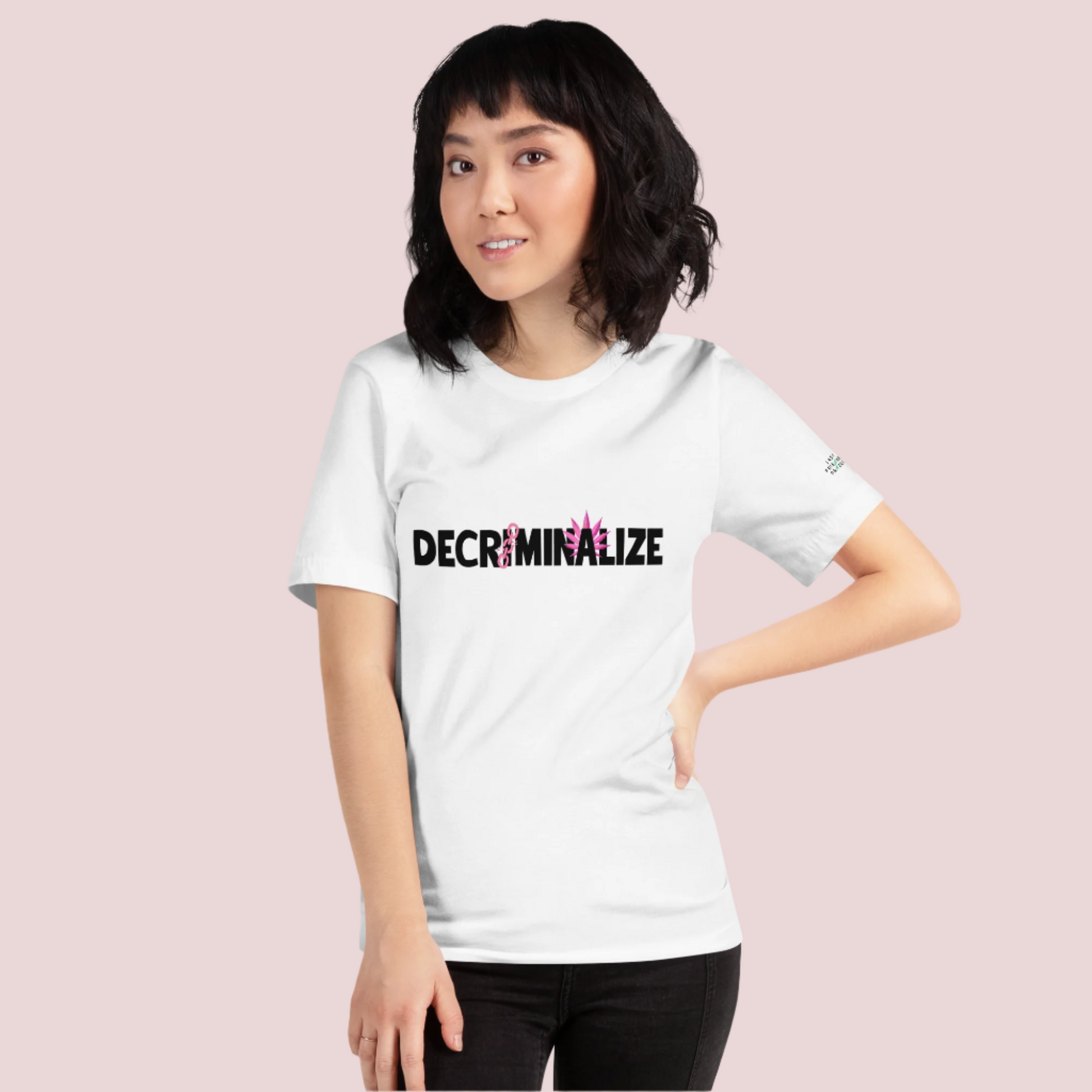 A person wearing a white crewneck tee showing the text "DECRIMINALIZE", with a broken pink chain over the first letter "I" and pink cannabis leaves behind the letter "A" . The Last Prisoner Project x The Counselle Collection logo on the left sleeve.