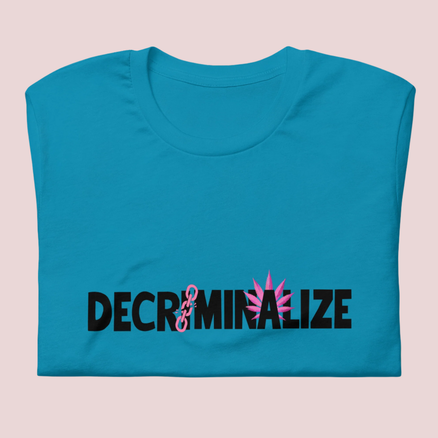 A folded blue crewneck tee showing the text "DECRIMINALIZE", with a broken pink chain over the first letter "I" and pink cannabis leaves behind the letter "A" .