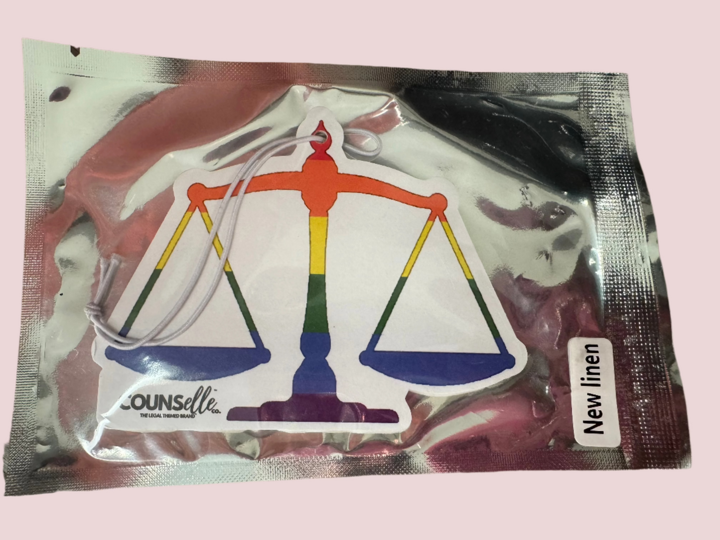 A picture of a car freshener in silver packaging. The freshener is in the shape of a justice scale and features the counselle collection® logo.
