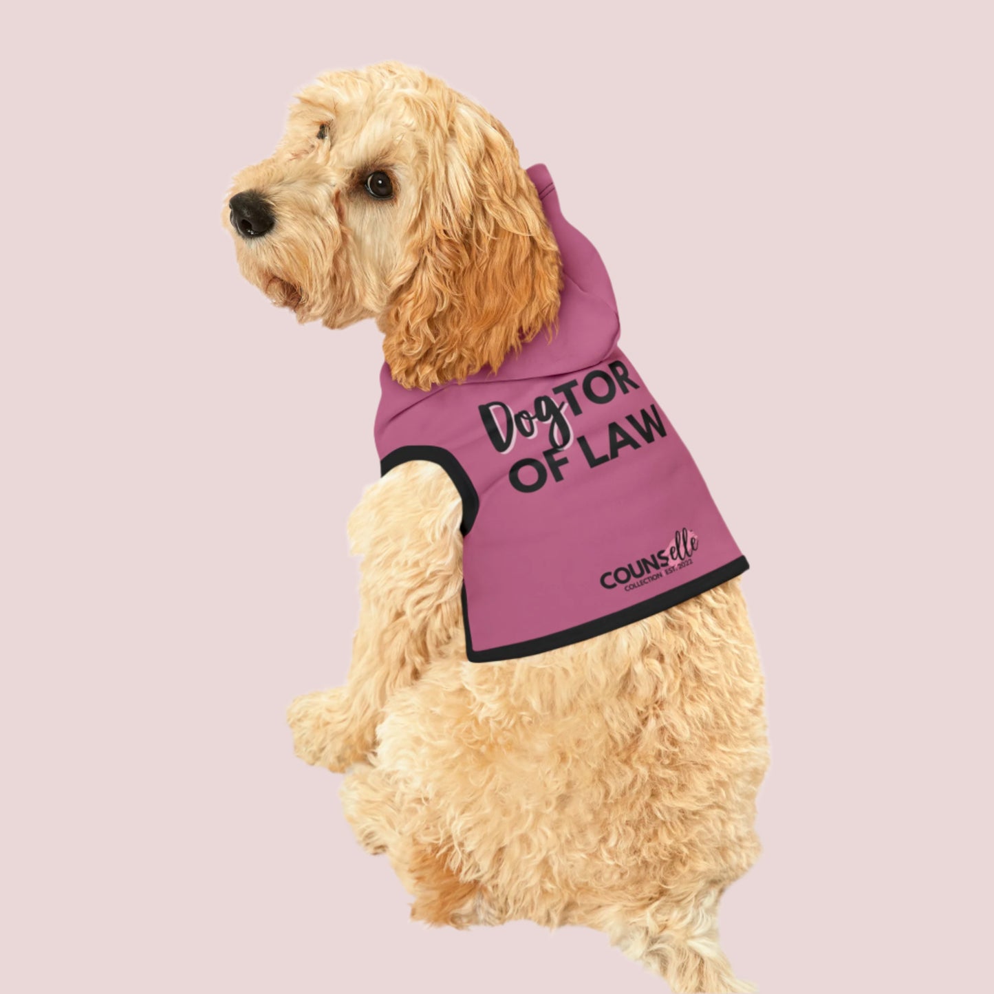 A dog wearing a pink pet hoodie with black cuffs and hem accents, featuring the words "DogTOR OF LAW" and the counsellecollection logo near the hemline.