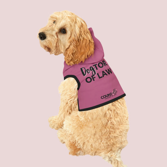 A dog wearing a pink pet hoodie with black cuffs and hem accents,  featuring the words "DogTOR OF LAW" and the counsellecollection logo near the hemline.