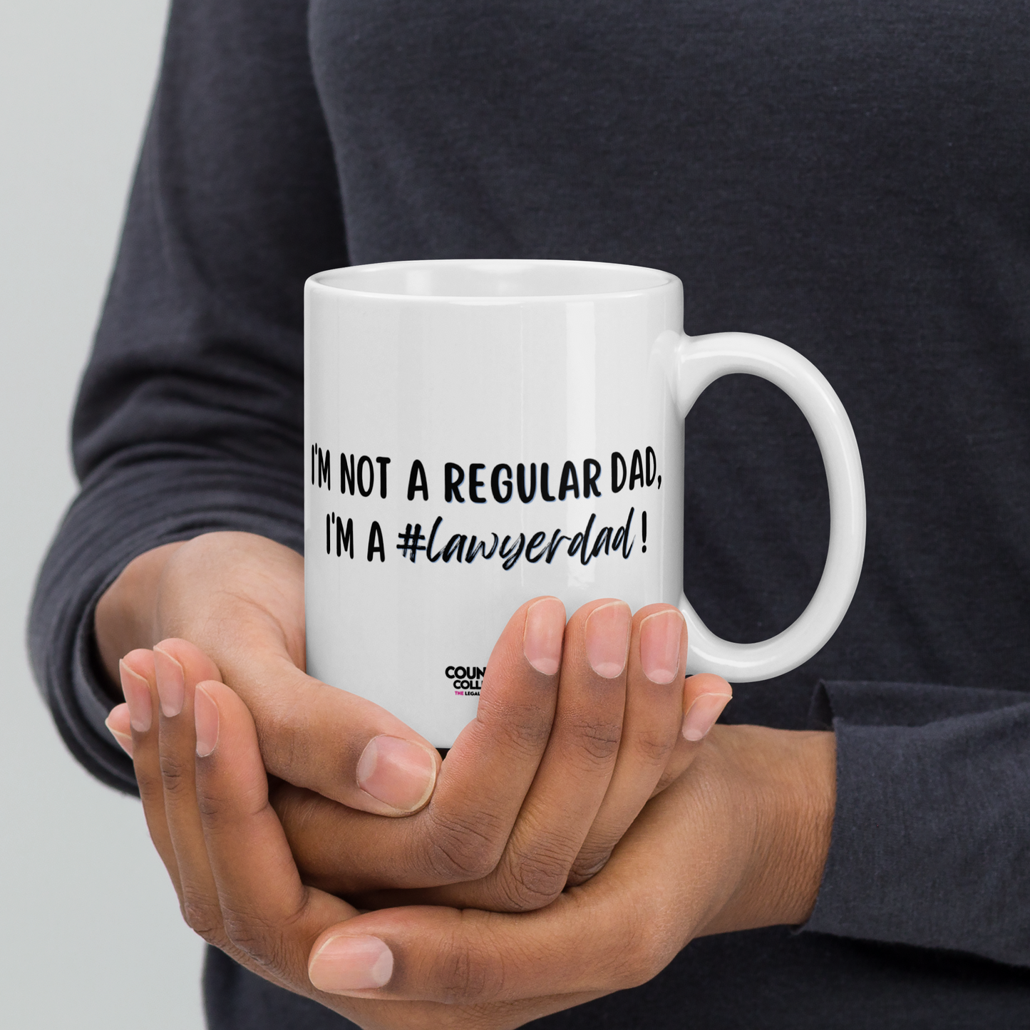 A model holding a white mug with the text "I'm not a regular dad, I'm a #lawyerdad!" and the counsellecollection logo at the bottom of the design.