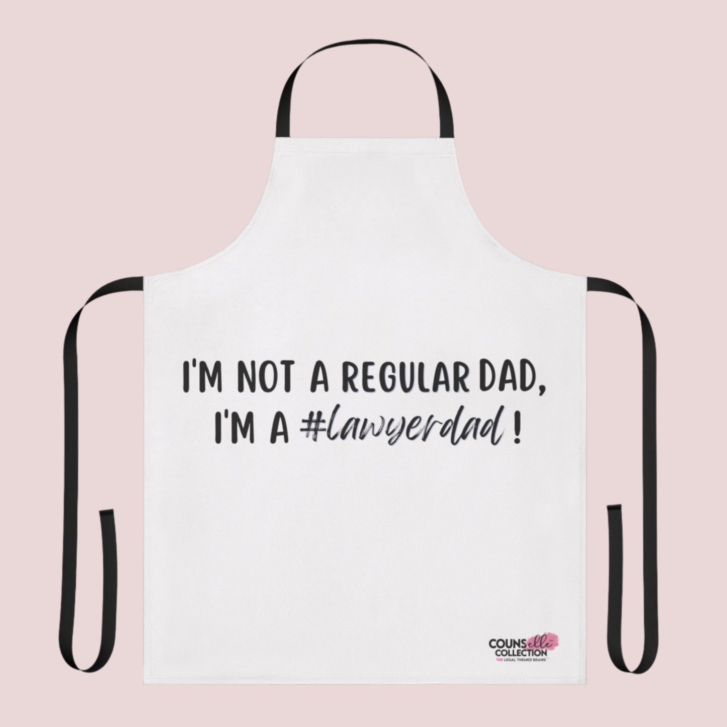 A white apron with the text "I'm not a regular dad, I'm a #lawyerdad!" and the counsellecollection logo on the bottom right corner. The apron has a black neck strap and belt.
