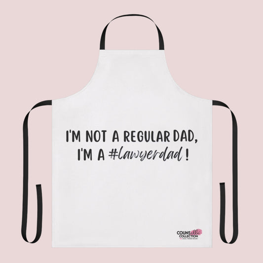A white apron with the text "I'm not a regular dad, I'm a #lawyerdad!"  and the counsellecollection logo on the bottom right corner. The apron has a black neck strap and belt.