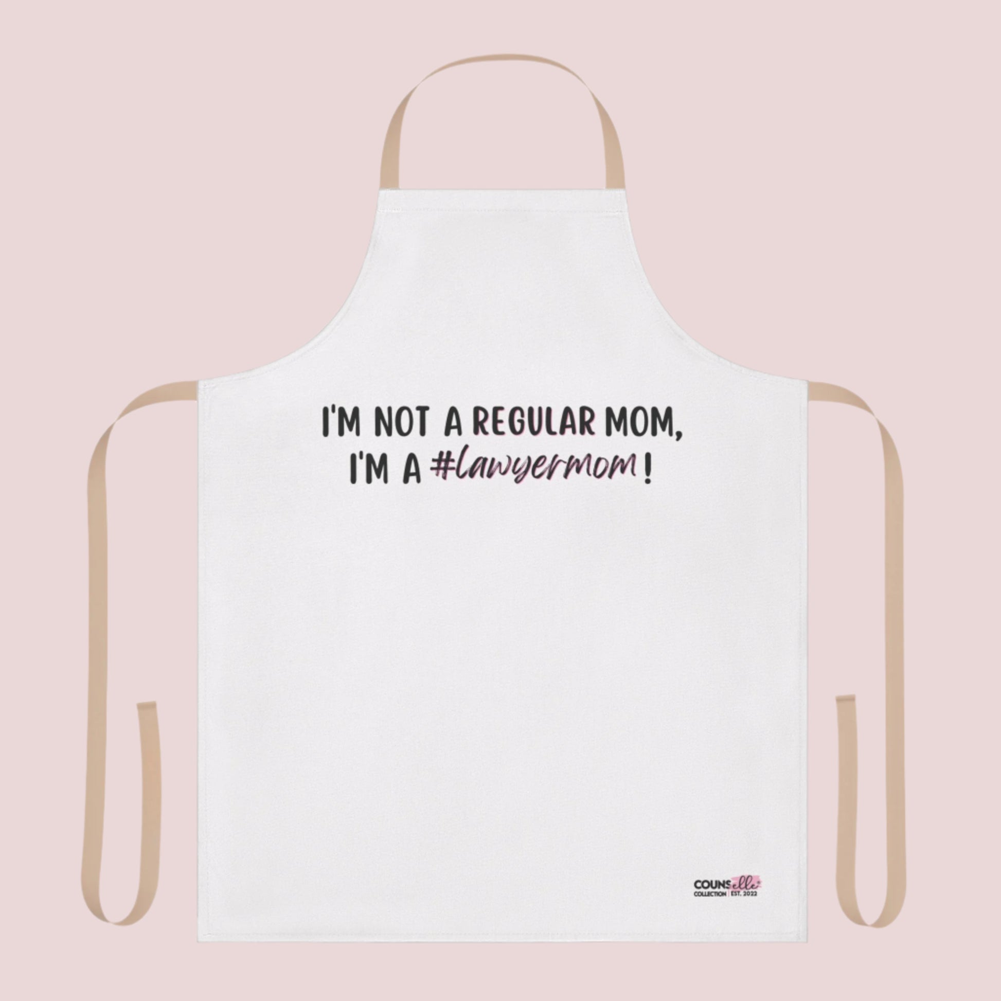 A white apron with the text "I'm not a regular mom, I'm a #lawyermom!"  and the counsellecollection logo on the bottom right corner. The apron has a neutral/beige strap and belt.