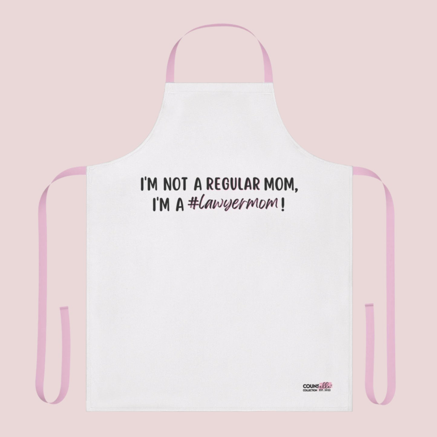 A white apron with the text "I'm not a regular mom, I'm a #lawyermom!" and the counsellecollection logo on the bottom right corner. The apron has a pink neck strap and pink belt.