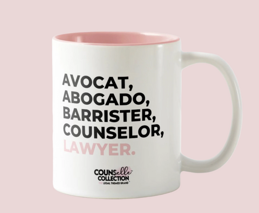 A white mug with a blush pink rim and inside color, with the text "avocat", "abogado", "barrister", "counselor", "legale", and "lawyer" on the front. Counselle Collection® logo under text.