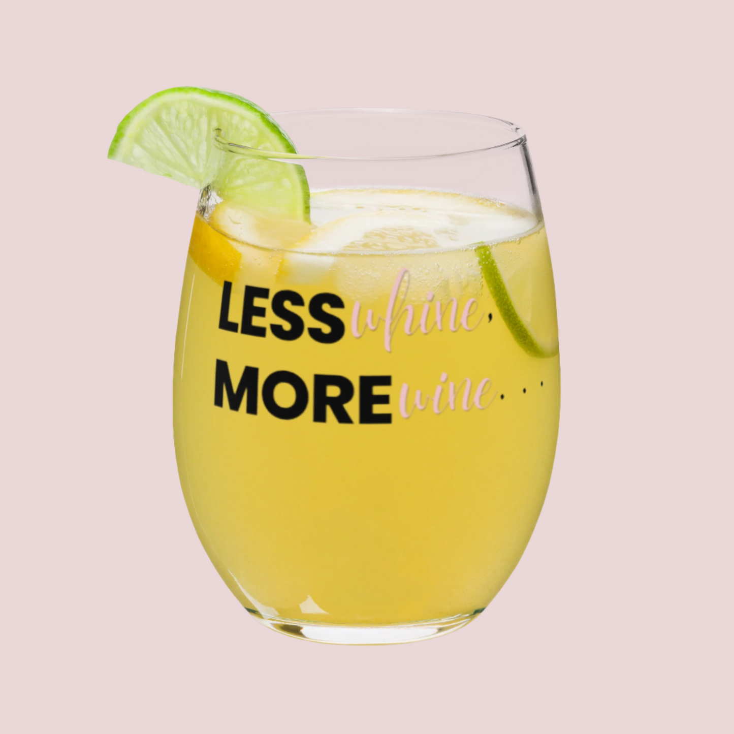 A clear, stemless wine glass with the words "Less WHINE, More WINE" written on it. Filled with a cocktail and lime slices, with a lime slice on the rim.