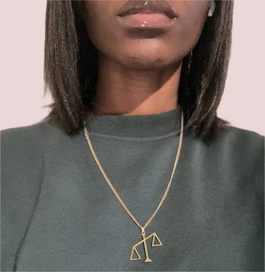 A gold chain necklace featuring a pendant in the shape of the scales of justice tipped to one side.