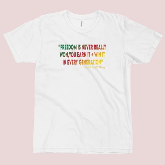 A white roundneck tee featuring the quote "FREEDOM IS NEVER REALLY WON, YOU EARN IT + WIN IT In EVERY GENERATION" followed by the text "-Carlotta Scott King", all are written in green, red, and yellow striped font.