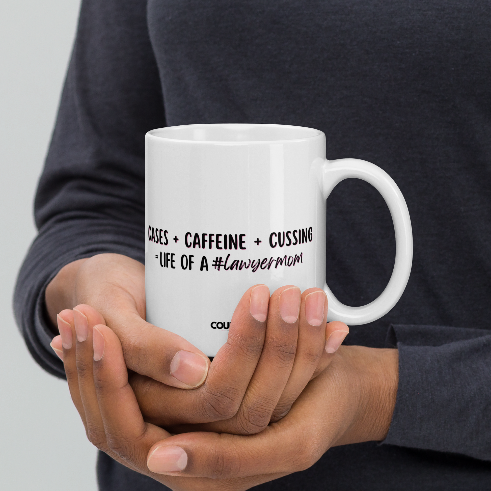 A person holding an 11oz white mug with the text "Cases + Caffeine + Cussing = LIFE OF A #lawyermom", and the counsellecollection logo at the bottom of the design