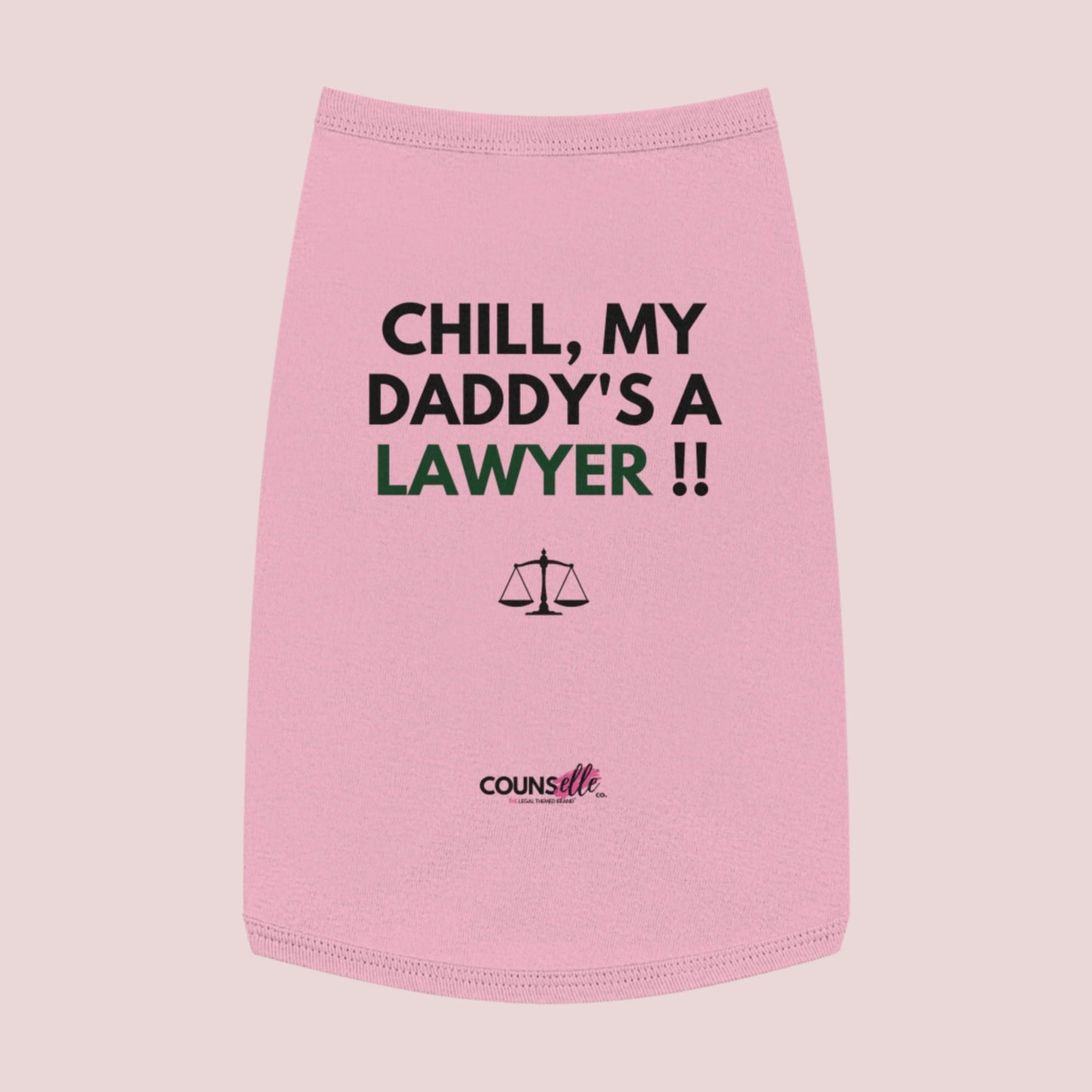 A pink pet tee featuring the quote "Chill, my daddy's a lawyer" in uppercase text, an illustration of justice scales below the text and the counsellecollection logo near the hem.