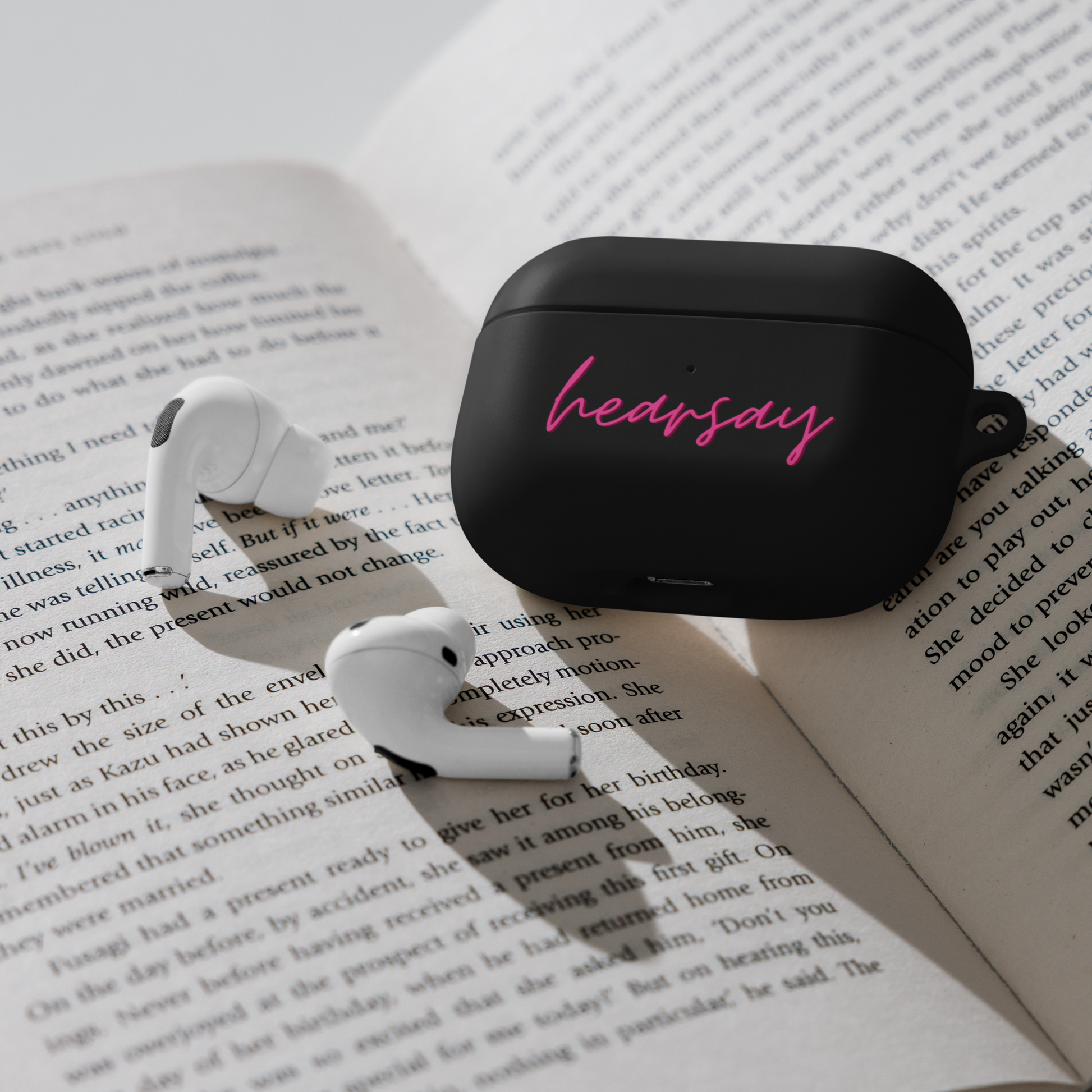 A black AirPods Case showing the word "hearsay" in script font laying flat inside an open book. Two airpods can also be seen beside the case.