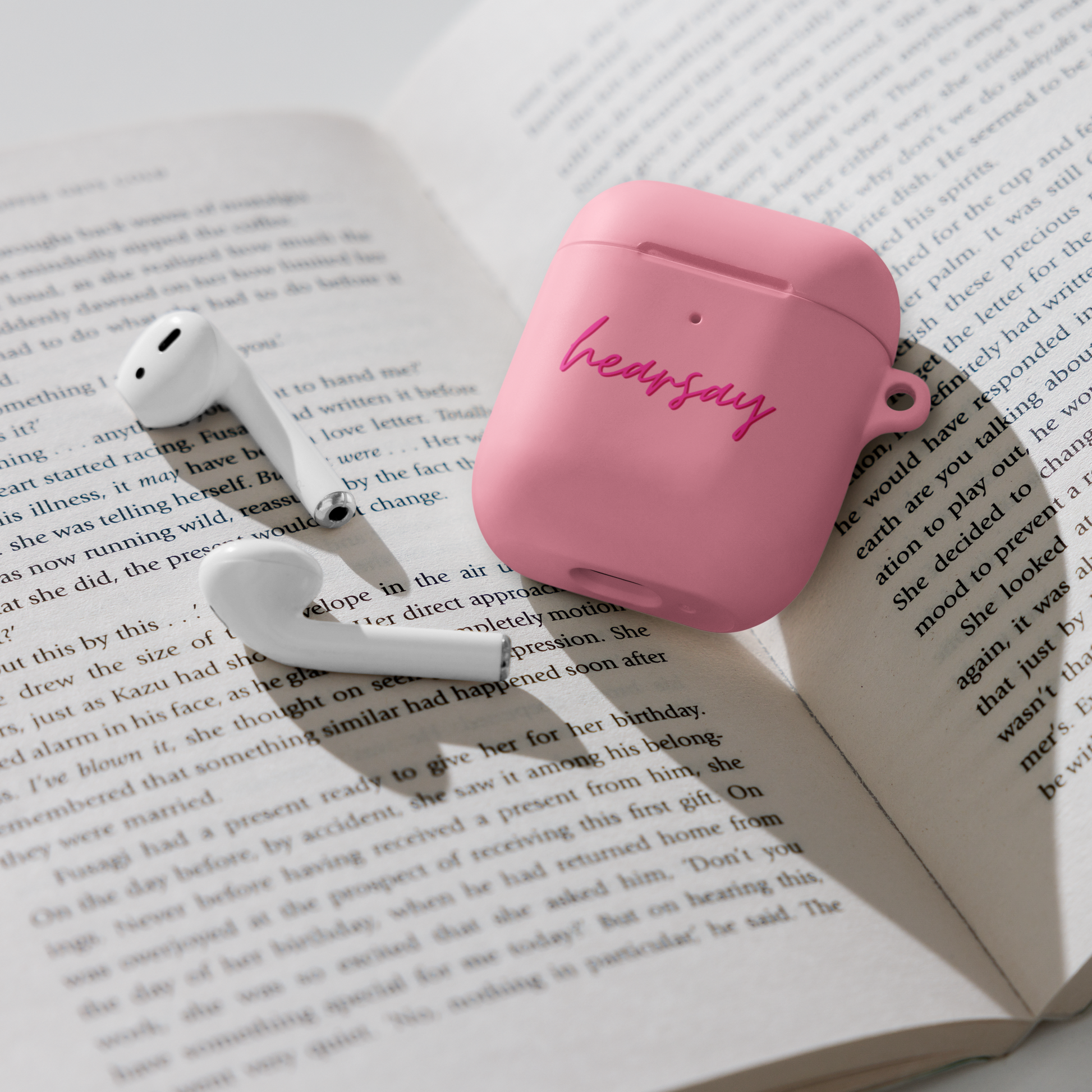 A pink AirPods Case showing the word "hearsay" in script font laying flat inside an open book. Two airpods can also be seen beside the case.