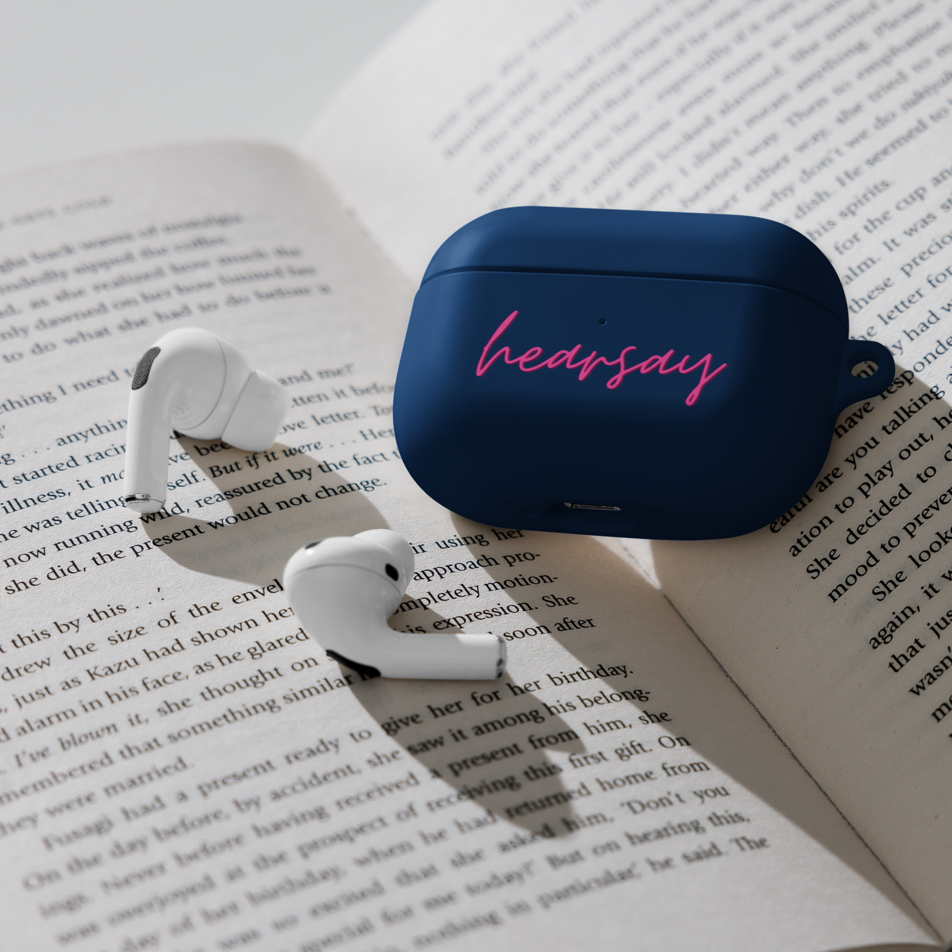 A navy blue AirPods Case showing the word "hearsay" in script font laying flat inside an open book. Two airpods can also be seen beside the case.
