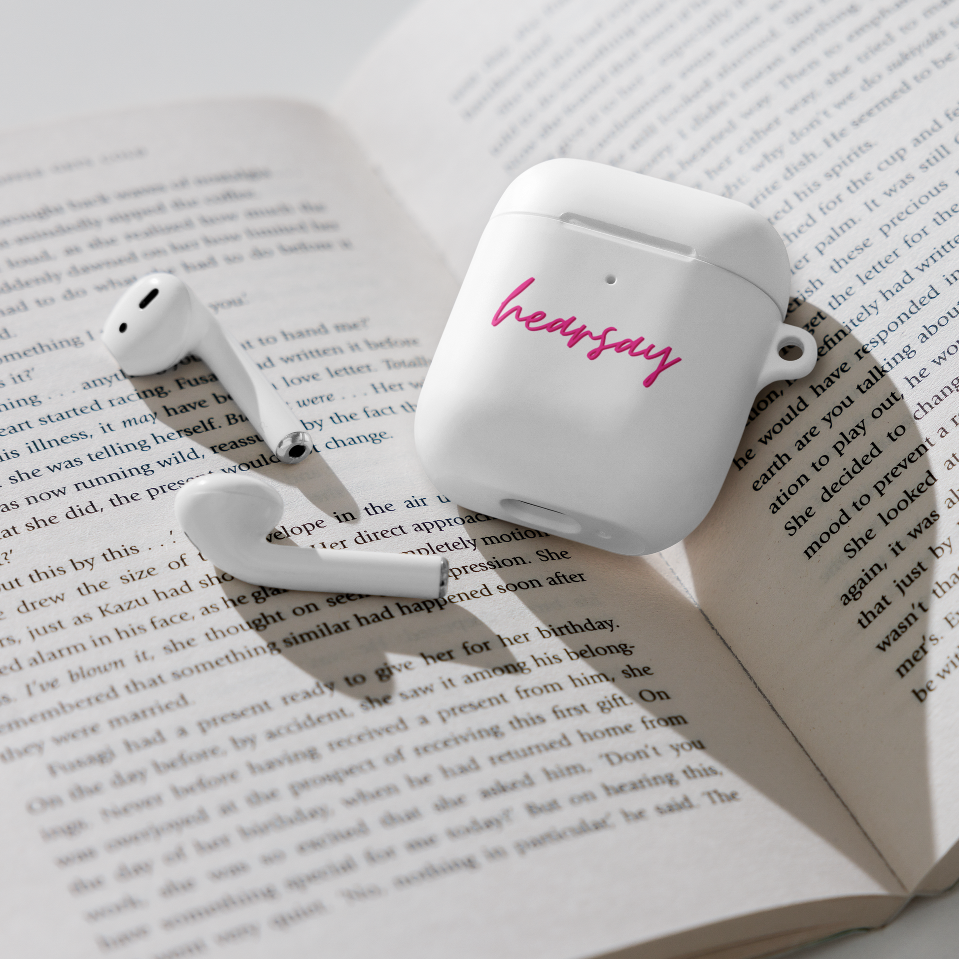 A white AirPods Case showing the word "hearsay" in script font laying flat inside an open book. Two airpods can also be seen beside the case.