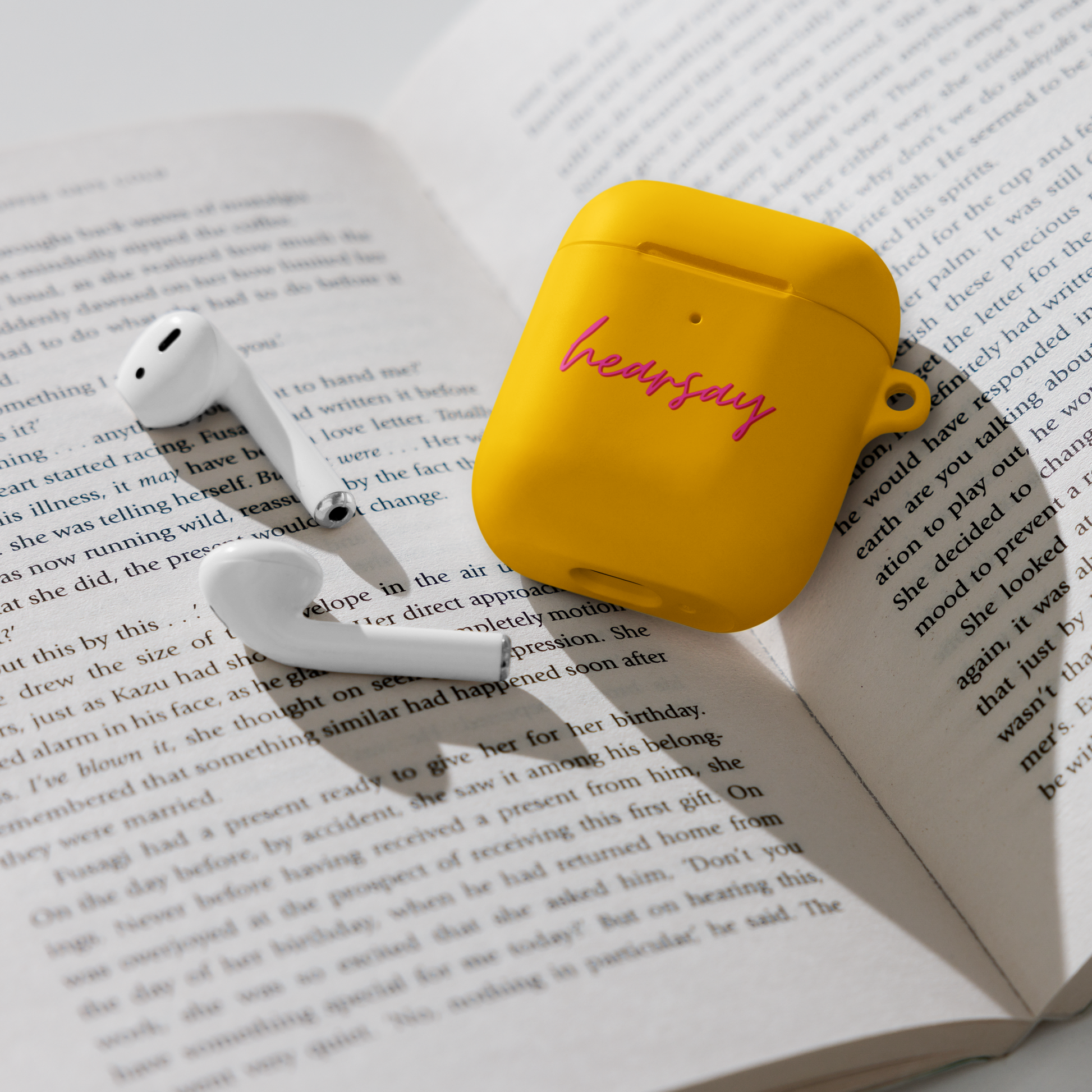 A yellow AirPods Case showing the word "hearsay" in script font laying flat inside an open book. Two airpods can also be seen beside the case.