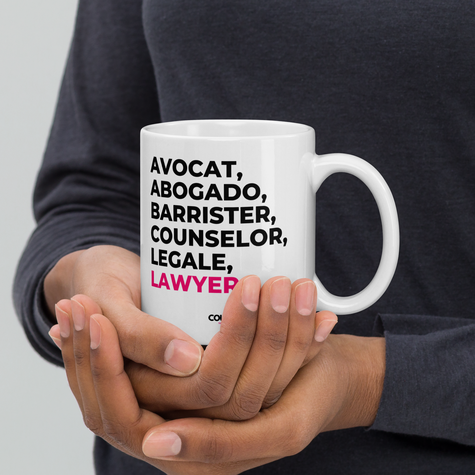A person in a dark grey shirt holding a white mug with the text "avocat", "abogado", "barrister", "counselor", "legale", and "lawyer" on the front. Counselle Collection® logo under text.