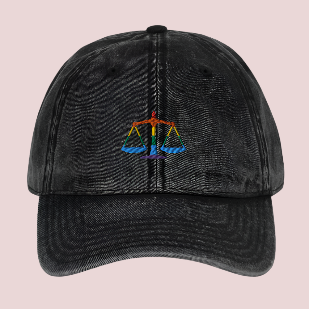 A black cap, featuring a justice scales in rainbow colors design.