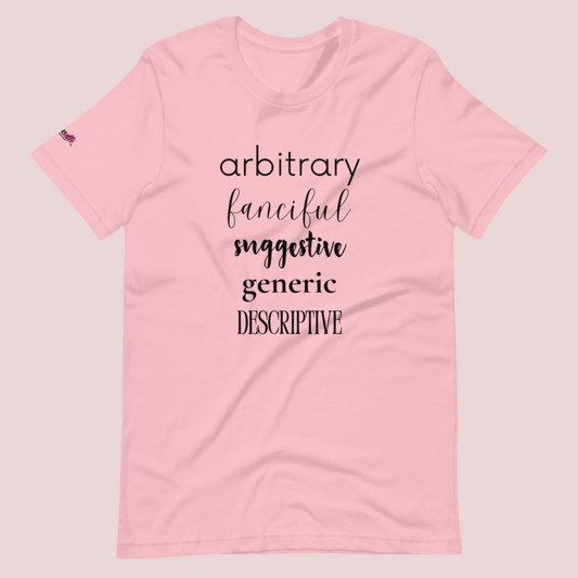 A pink roundneck tee featuring the words "arbitrary", "fanciful", "suggestive", "generic", and "descriptive" in different font styles, with the counsellecollection logo on a sleeve.