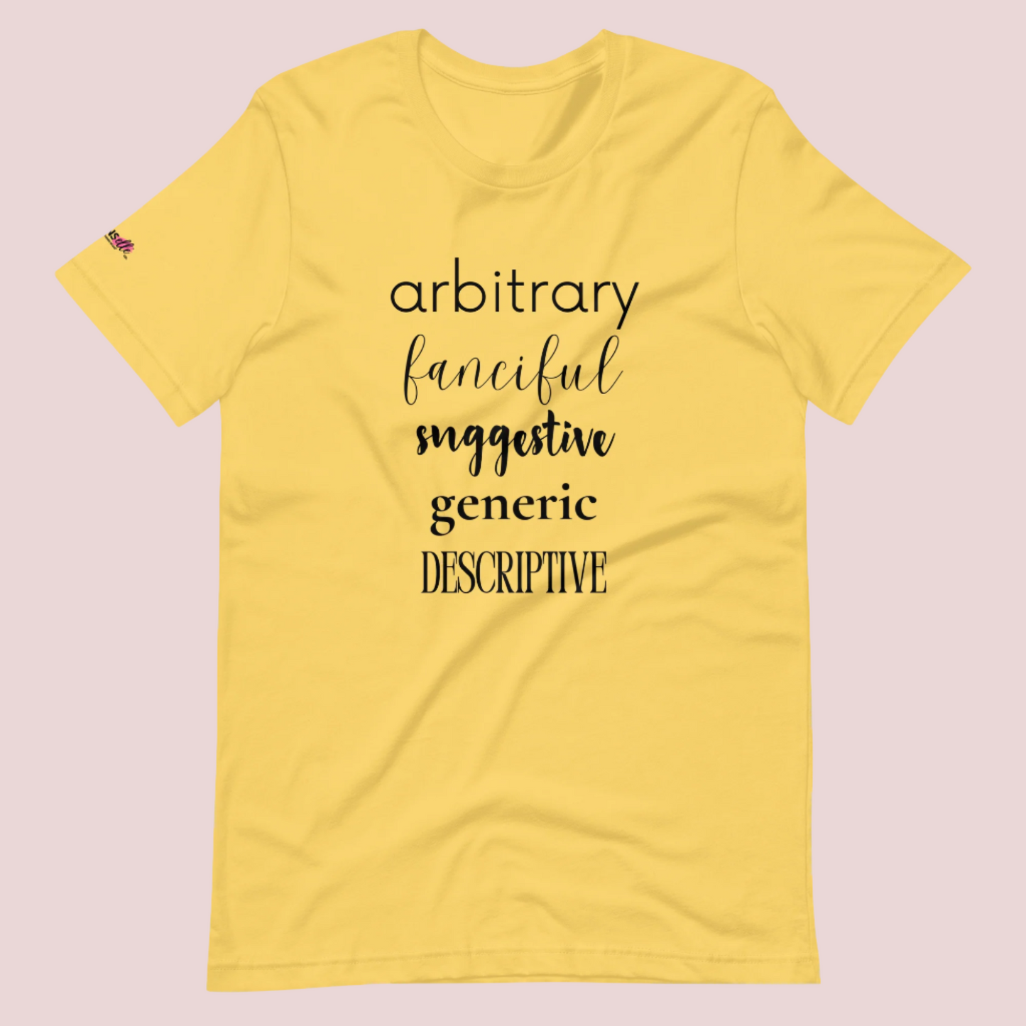 A yellow roundneck tee featuring the words "arbitrary", "fanciful", "suggestive", "generic", and "descriptive" in different font styles, with the counsellecollection logo on a sleeve.