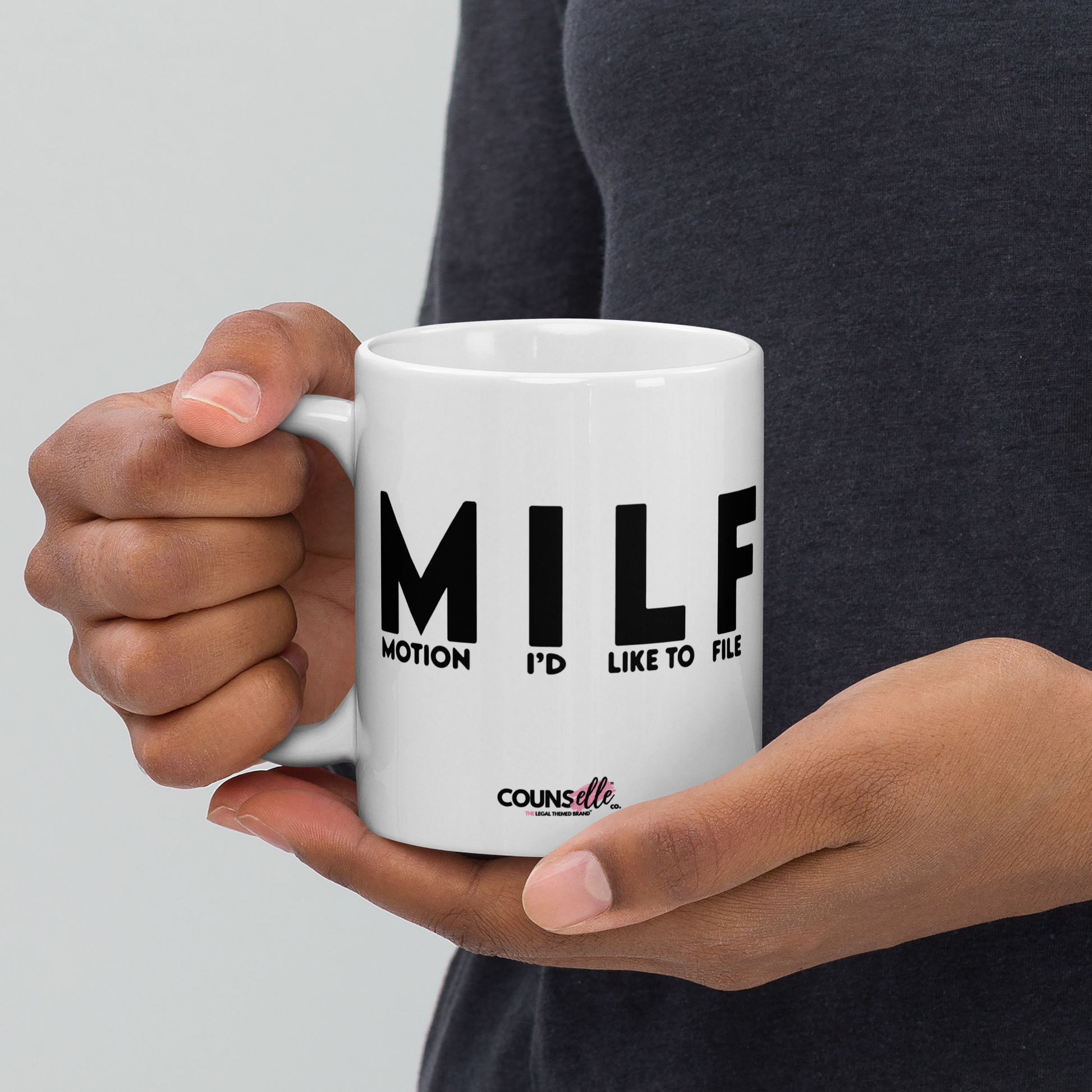 A person holding a white mug with the letters "MILF" and the words "MOTION I'D LIKE TO FILE" in black uppercase font and the counsellecollection logo at the bottom of the mug.