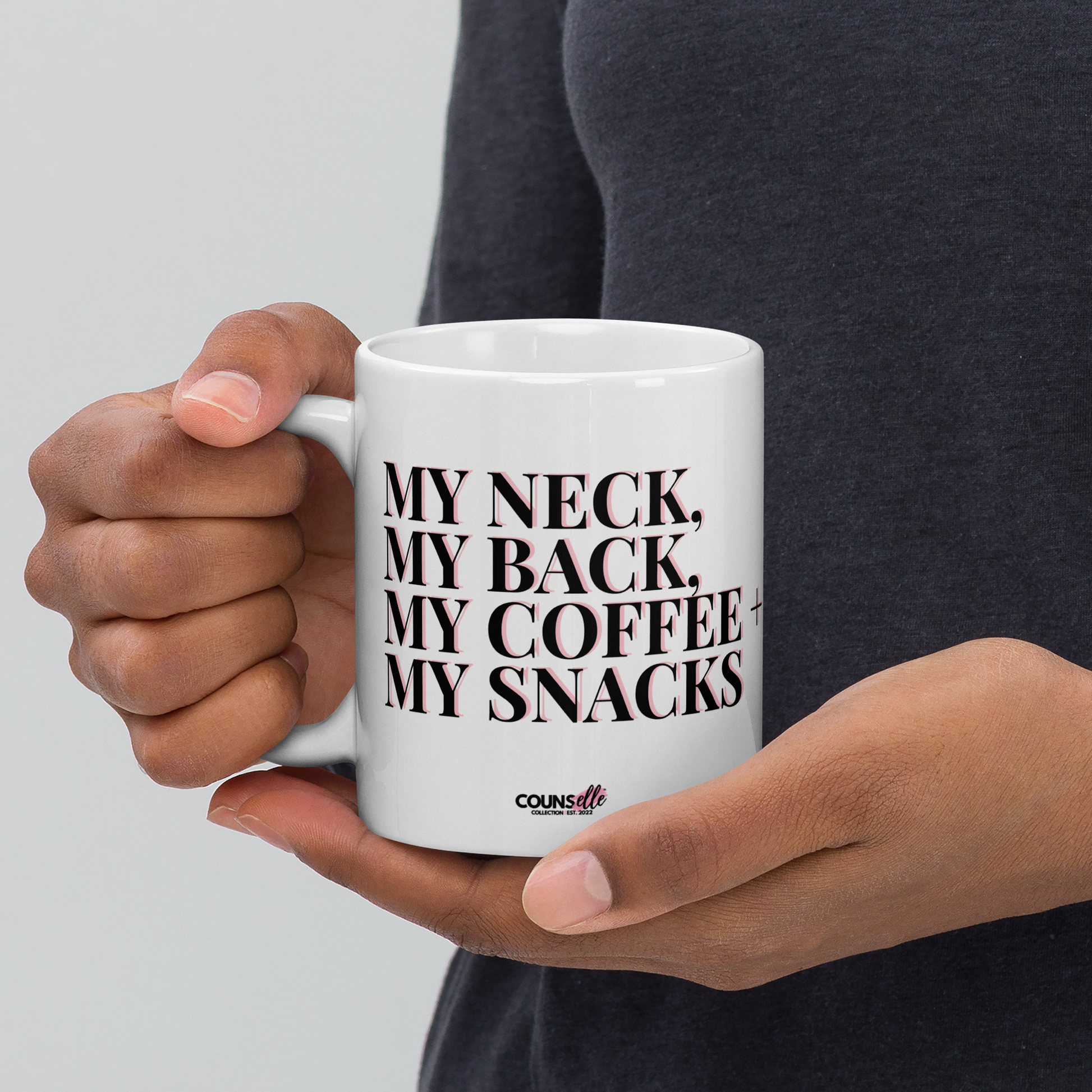 A person wearing a dark grey shirt holding a white ceramic mug featuring eye-catching bold block text: "MY NECK, MY BACK, MY COFFEE, MY SNACKS."