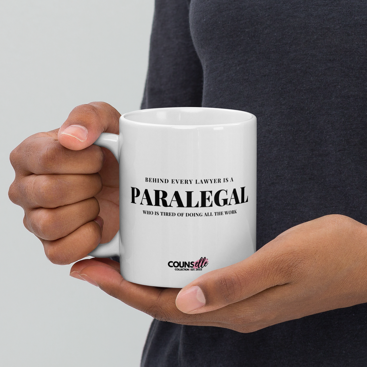 A person holding a white mug featuring the text "BEHIND EVERY LAWYER IS A PARALEGAL WHO IS TIRED OF DOING ALL THE WORK"