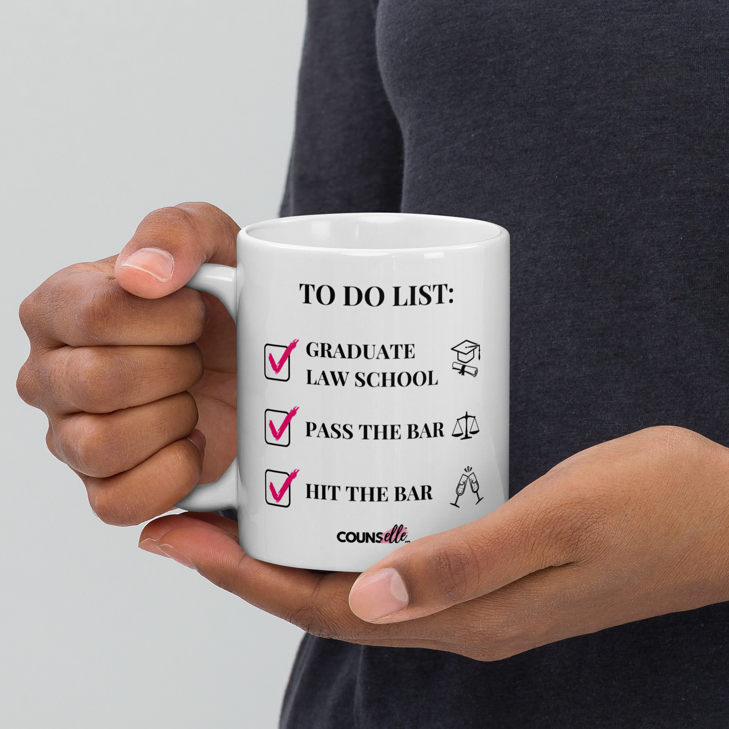A person holding a white mug showing the text "TO DO LIST", with ticked-off boxes beside the texts "GRADUATE LAW SCHOOL" and an illustration of a graduation cap and diploma; "PASS THE BAR" ans an illustration of justice scales; and "HIT THE BAR" and an illustration of clinking champagne flutes. The counsellecollection logo is seen at the bottom of the design.