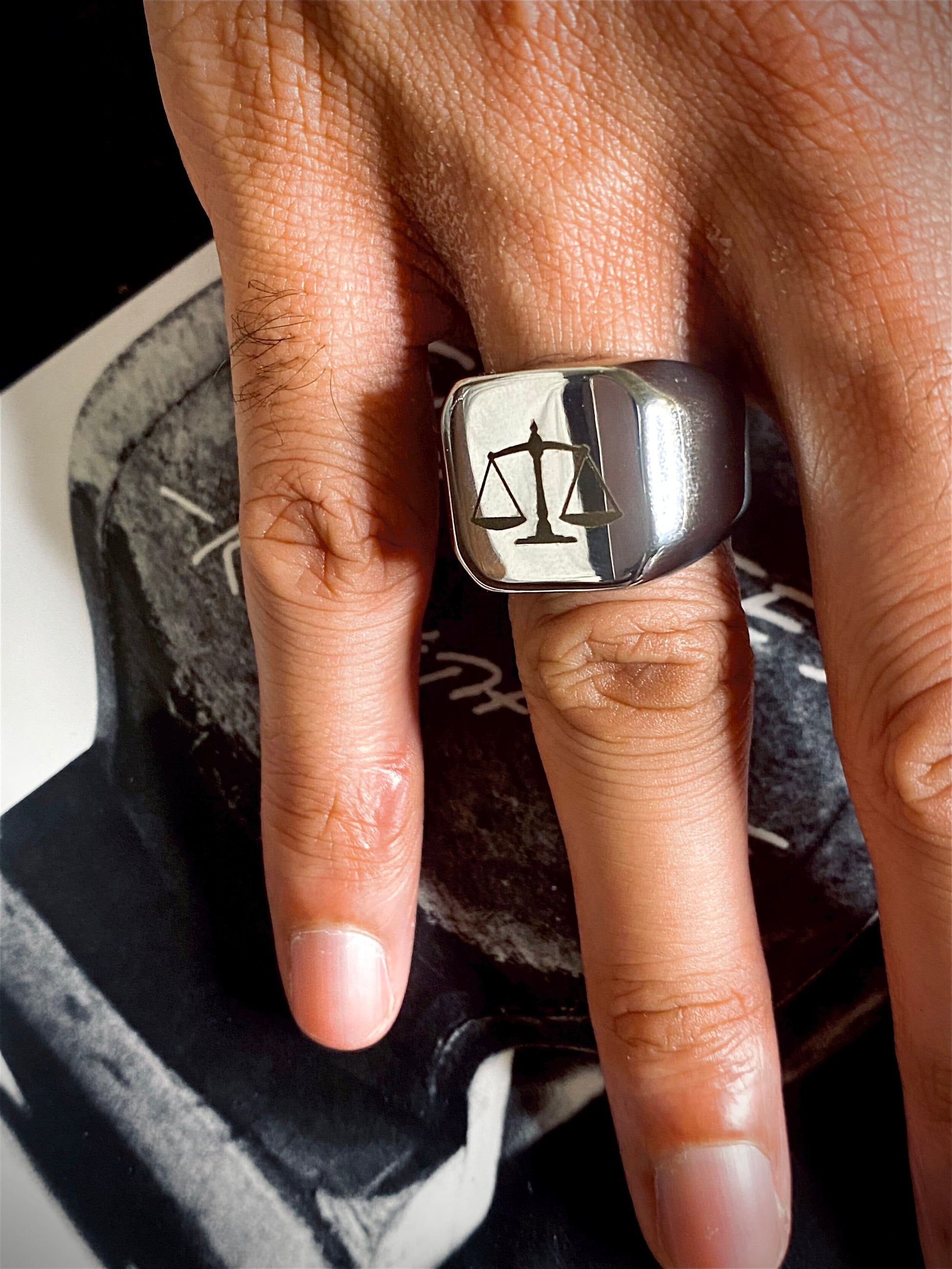 A sleek unisex ring featuring a justice scales emblem. Available in gold and silver.